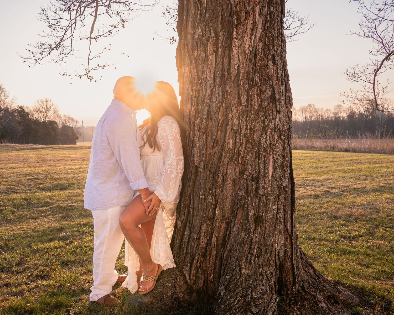Couple in white outfits sharing a romantic kiss against a large tree at golden hour in a scenic field.