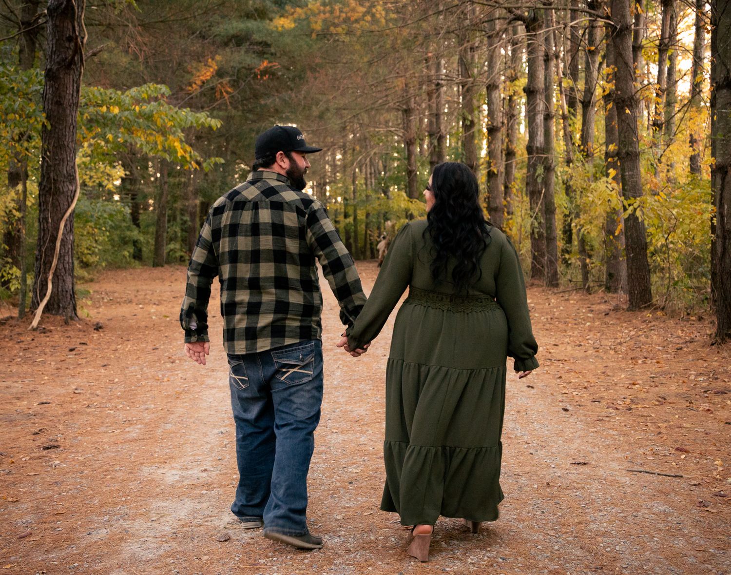 A couple walking hand in hand down a leaf-covered forest path during autumn with golden light filtering through trees.