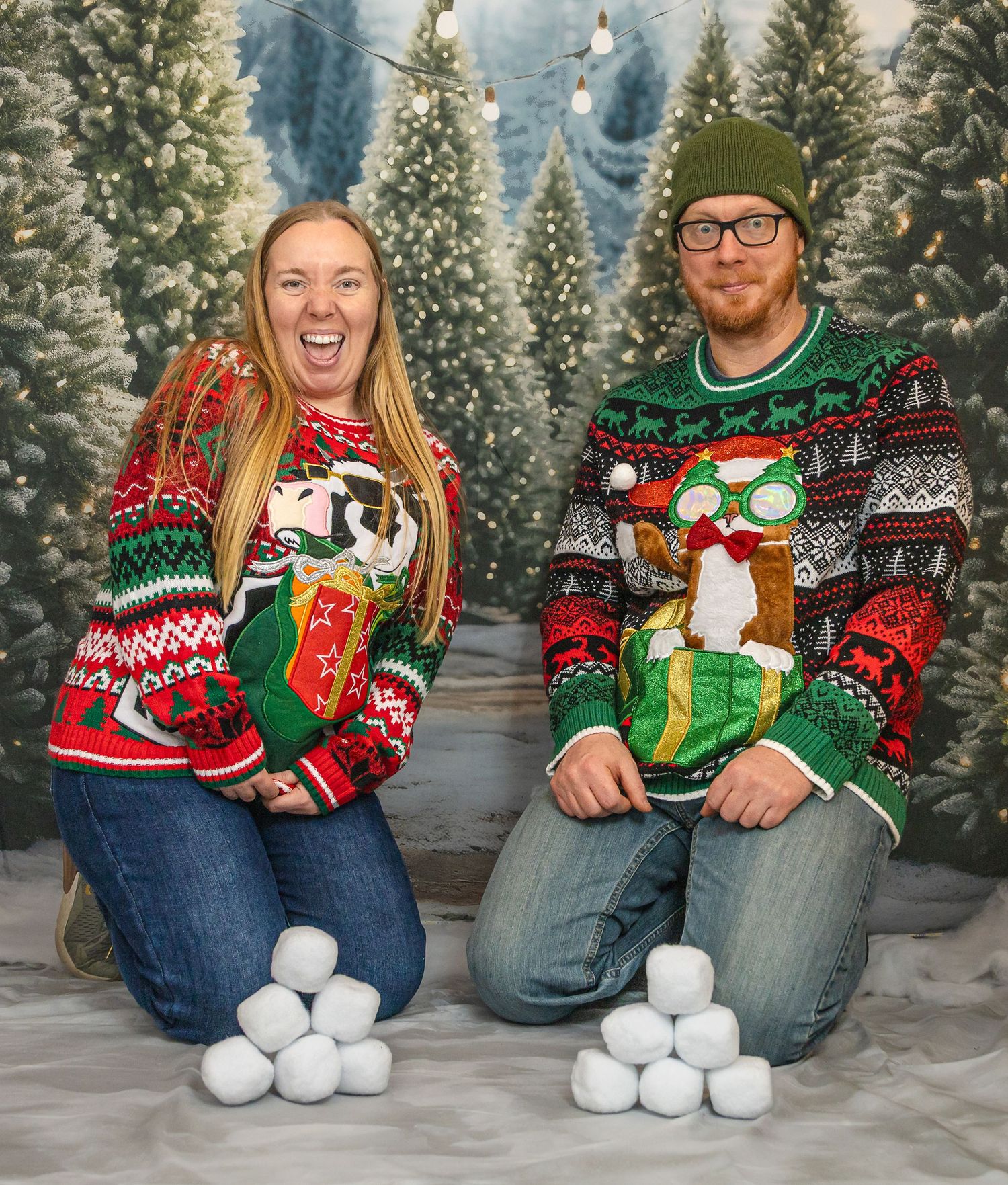 Couple in matching Christmas sweaters sitting with snowballs against a backdrop of evergreen trees and snow.
