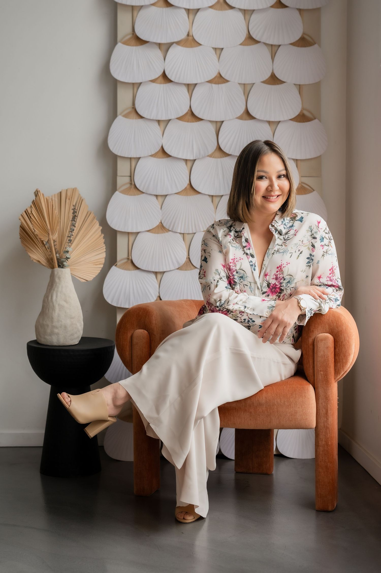 Stylish interior portrait featuring a floral blouse, cream pants, and modern decor with white scalloped wall art and dried palm.