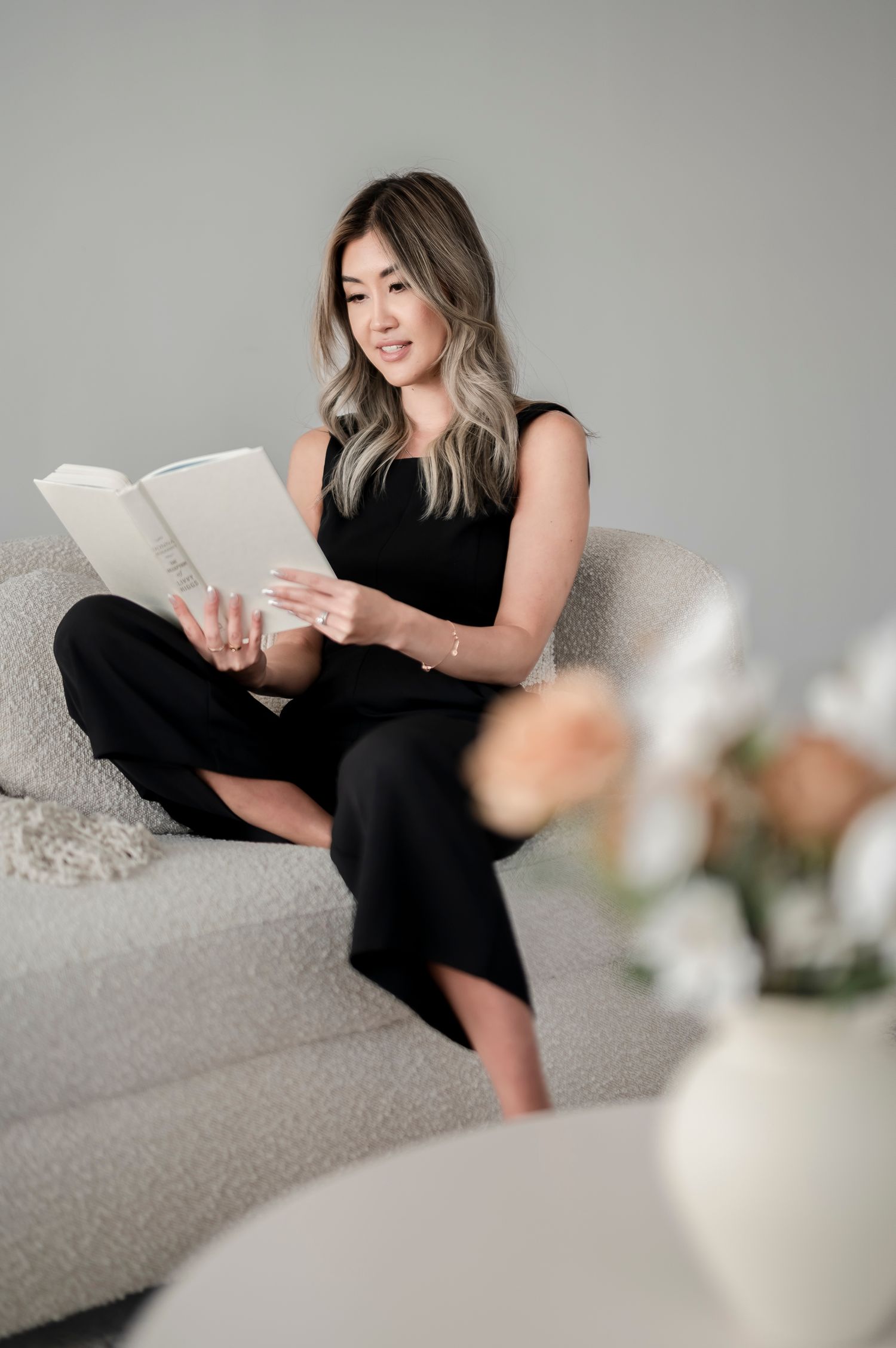 Person in black outfit relaxes on light gray couch while reading book, with white flowers in background.