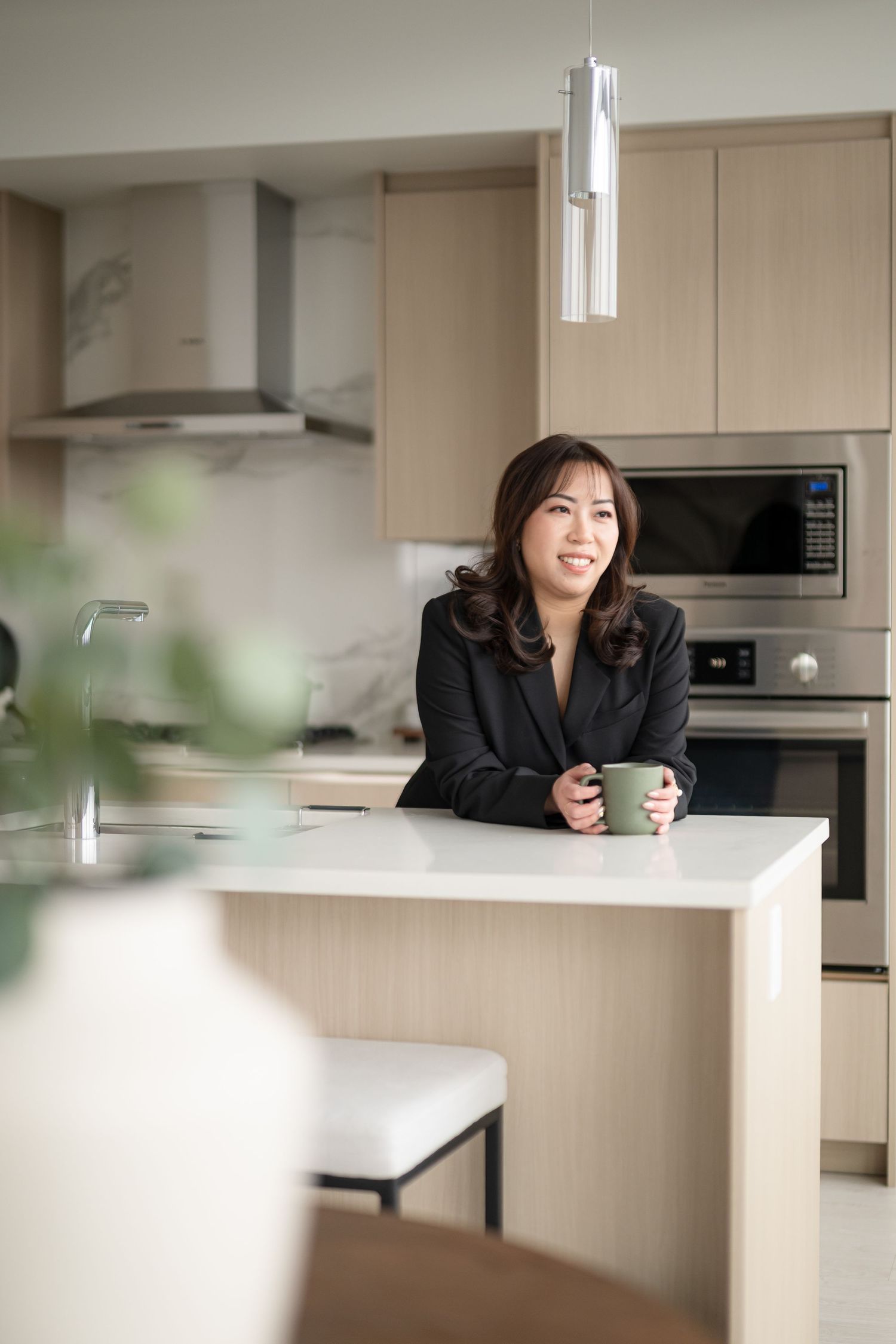 A person in a black blazer stands at a modern kitchen counter holding a coffee mug and smiling.