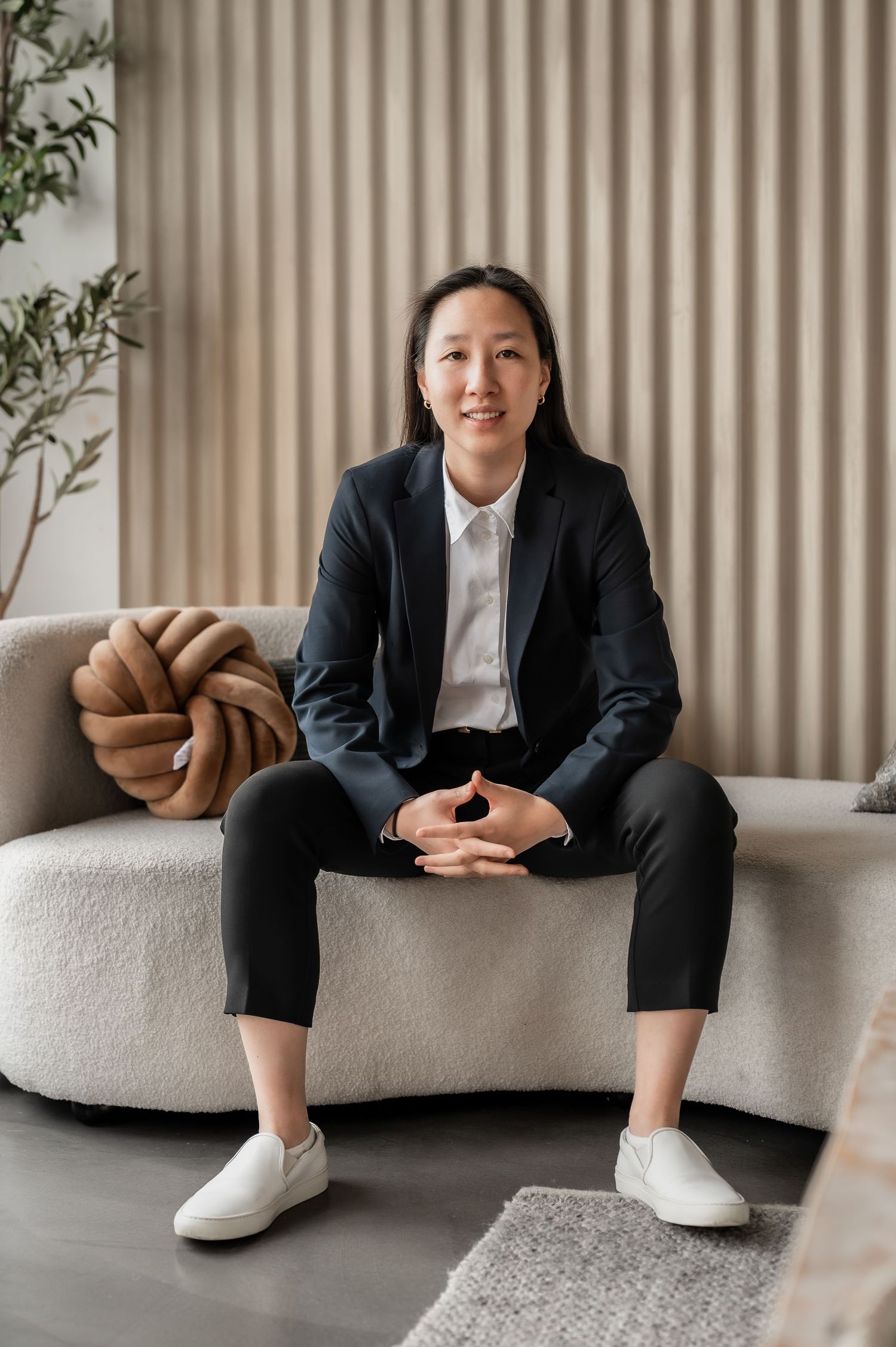 Professional in a navy blazer and white shirt sits casually on a beige couch with minimalist decor in the background.