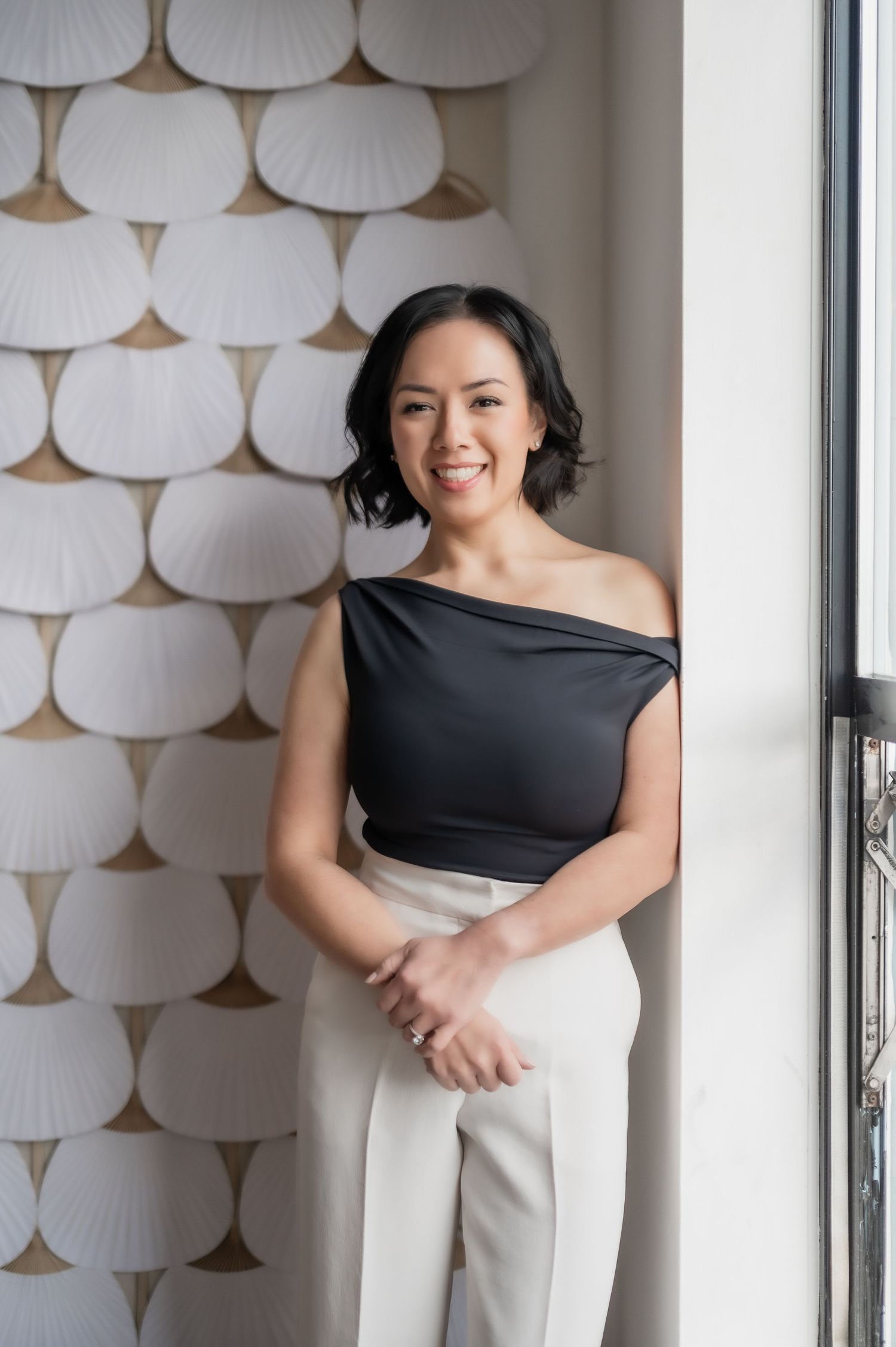 Professional portrait against white patterned wall featuring asymmetrical black top and white pants in modern setting.