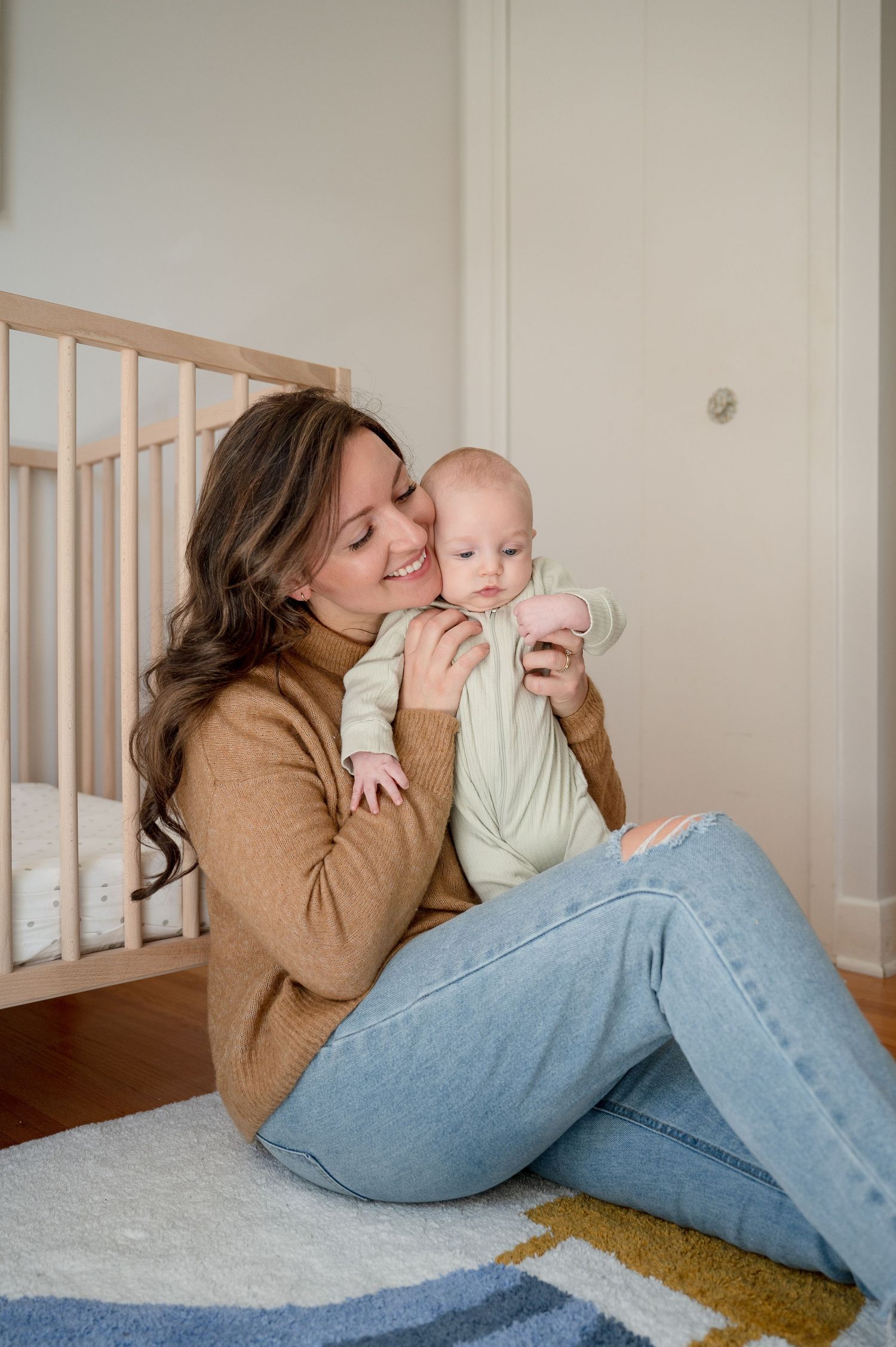 Caregiver in tan sweater and jeans sits on a floor rug by a crib while holding and cuddling with an infant in green.