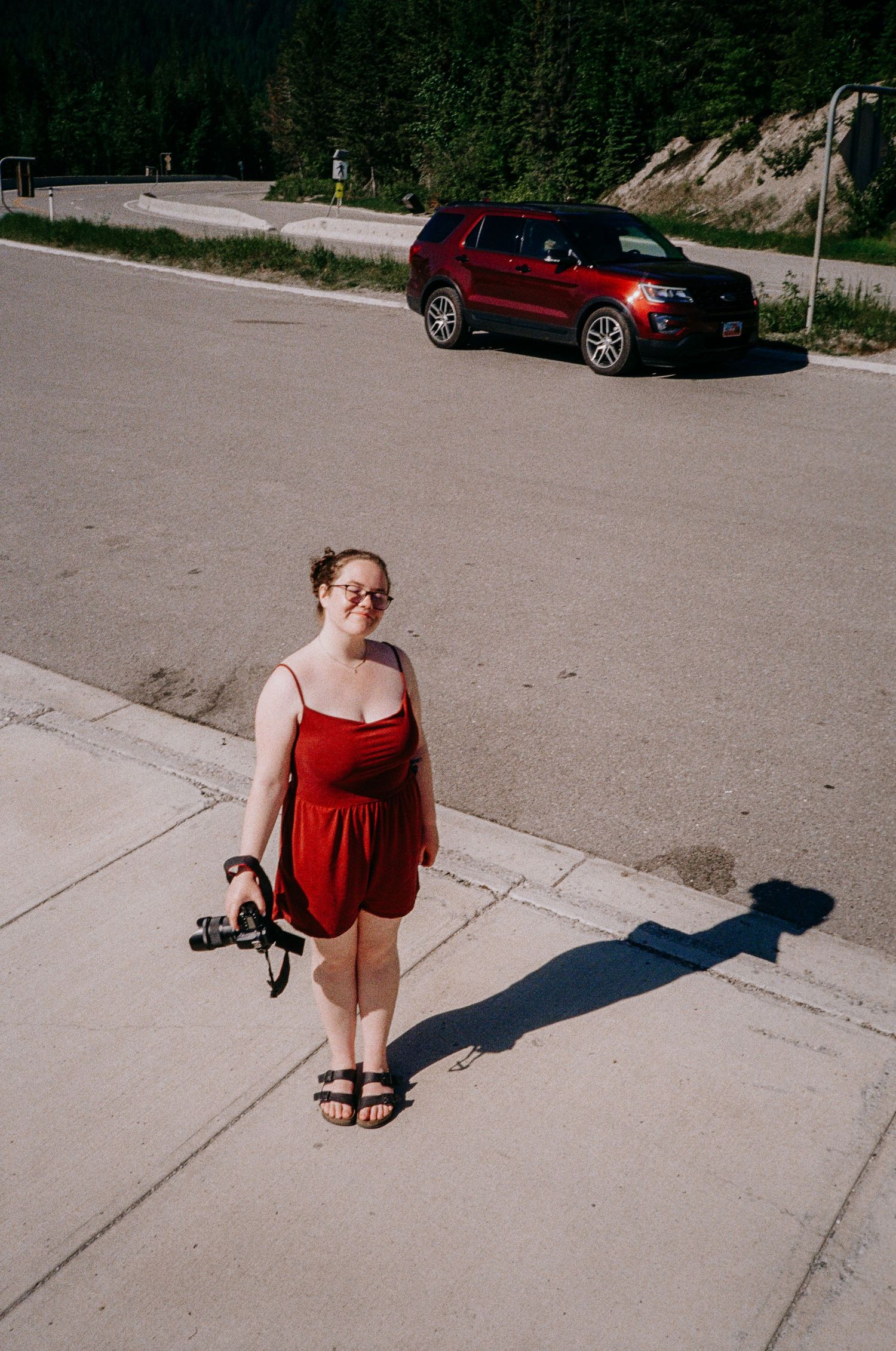 A person in red dress stands on sidewalk casting long shadow with car in background.