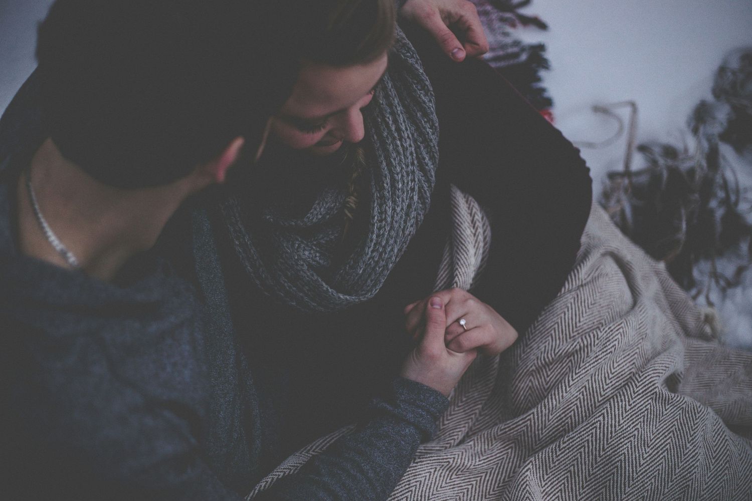 Couple holding hands intimately while sitting together under a cozy blanket in a dimly lit room.