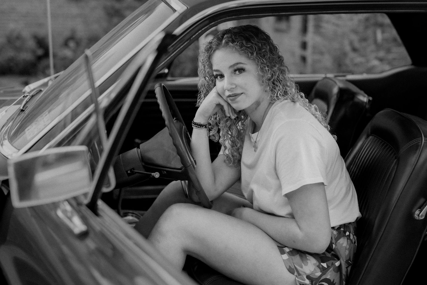 A young woman with curly hair leans forward, resting her chin on her hand and smiling softly in a vintage mustang.