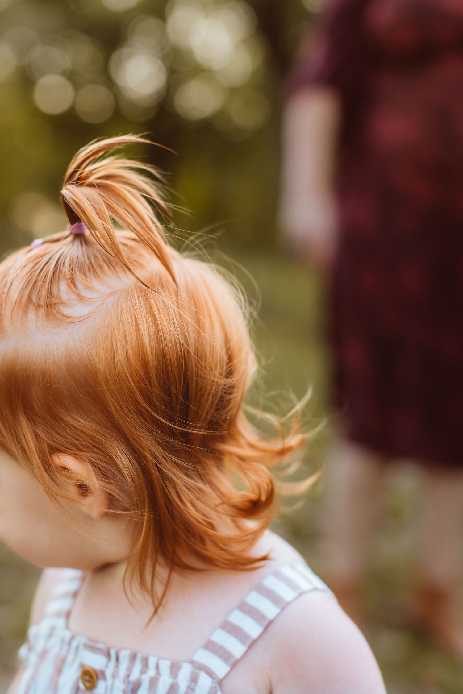 A toddler turns her face away so that it is not visible, bringing her red curls and whispy ponytail into focus.