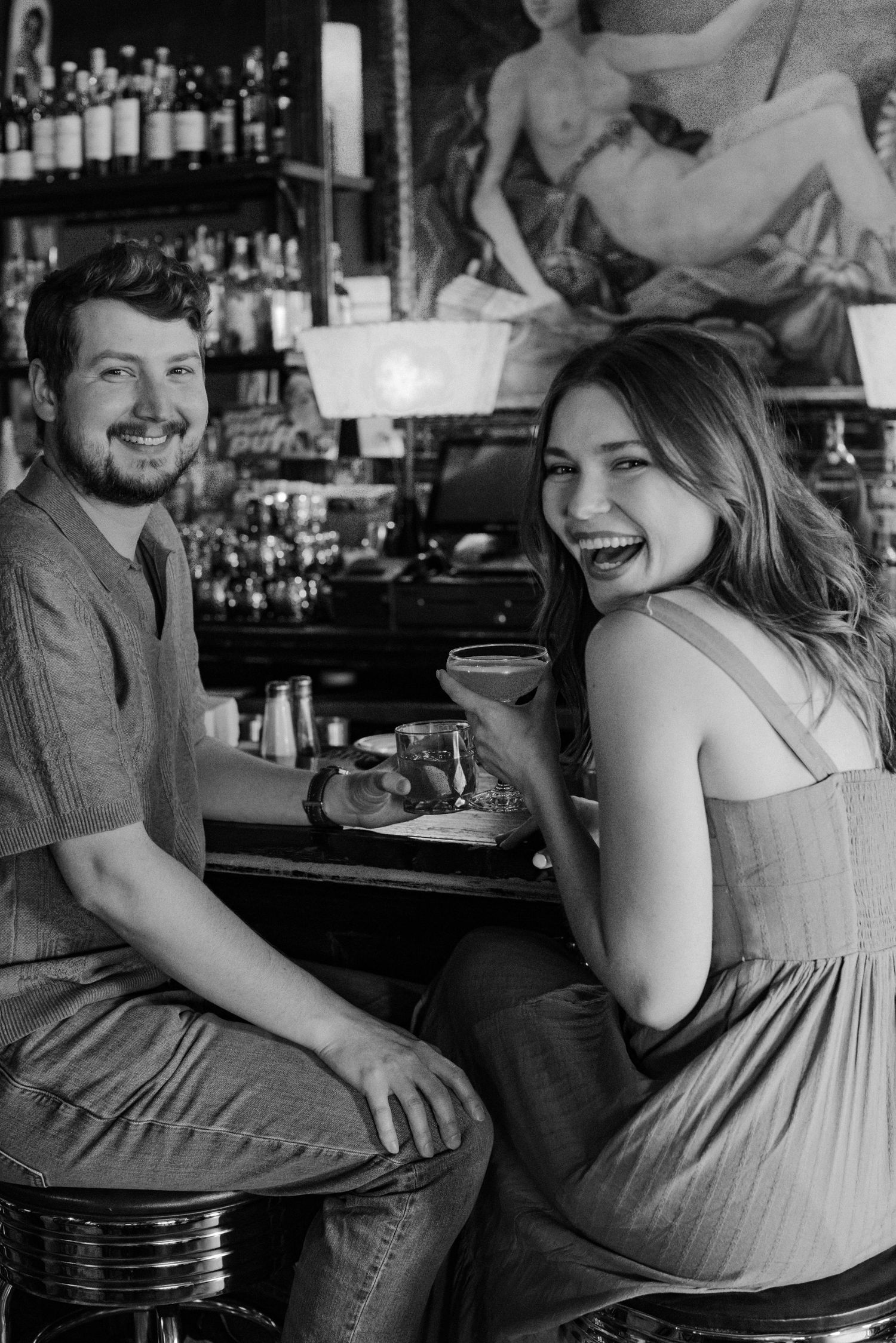 A black and white images of a woman holding a cocktail and laughing over her shoulder as her fiance smiles next to her at a bar.