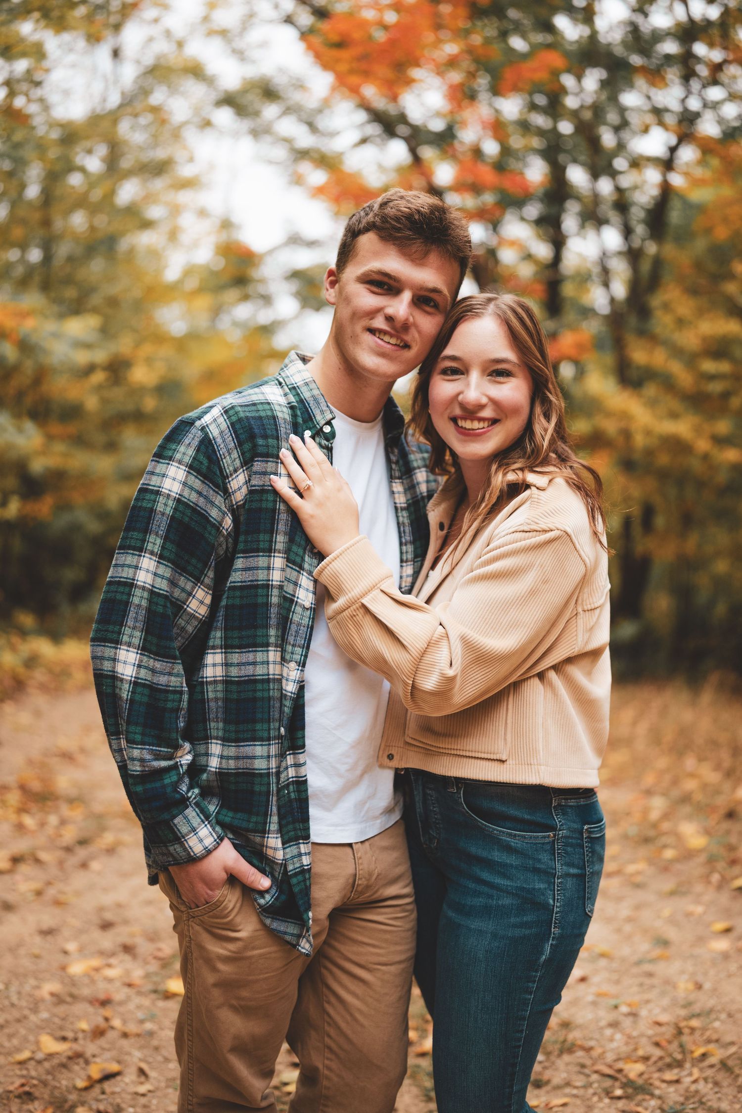 A happy couple poses together on a fall forest path, surrounded by orange and yellow autumn leaves.