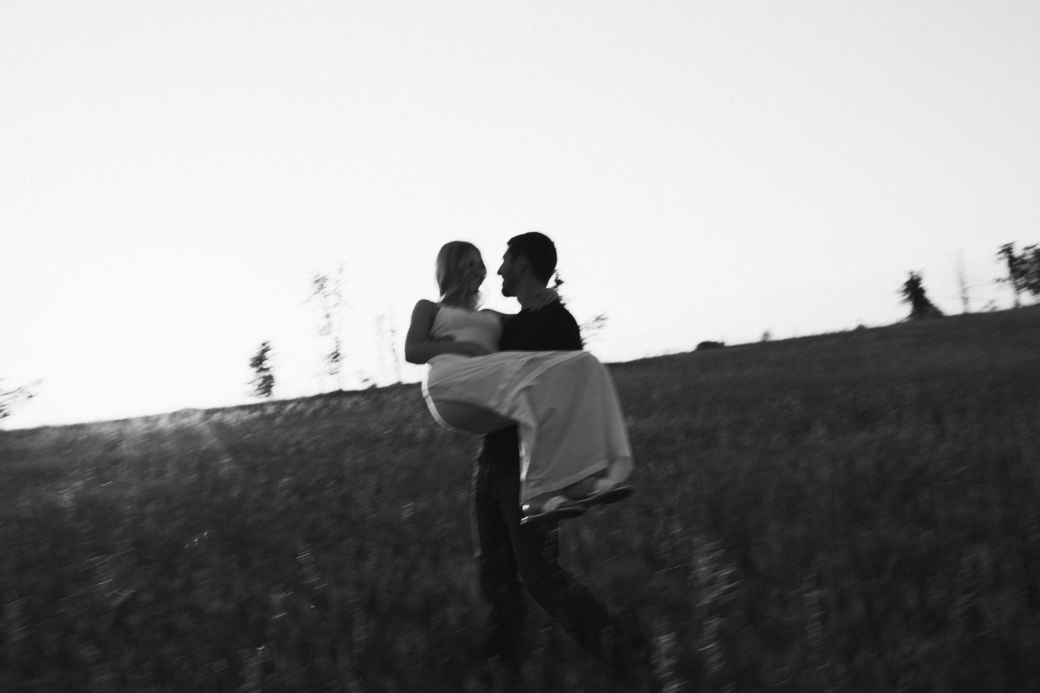 Black and white silhouette of man lifting woman in his arms on open hillside in soft fading light.