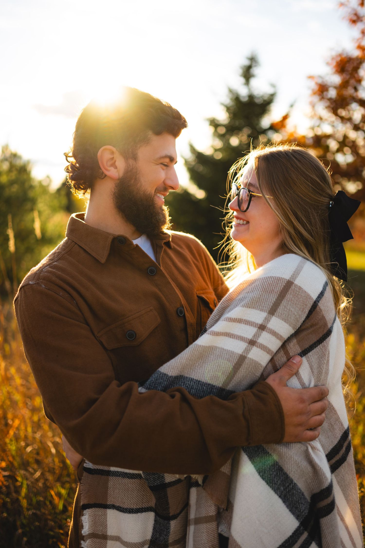 Couple embracing in golden sunset light, man in brown jacket smiling at woman wrapped in plaid blanket outdoors.