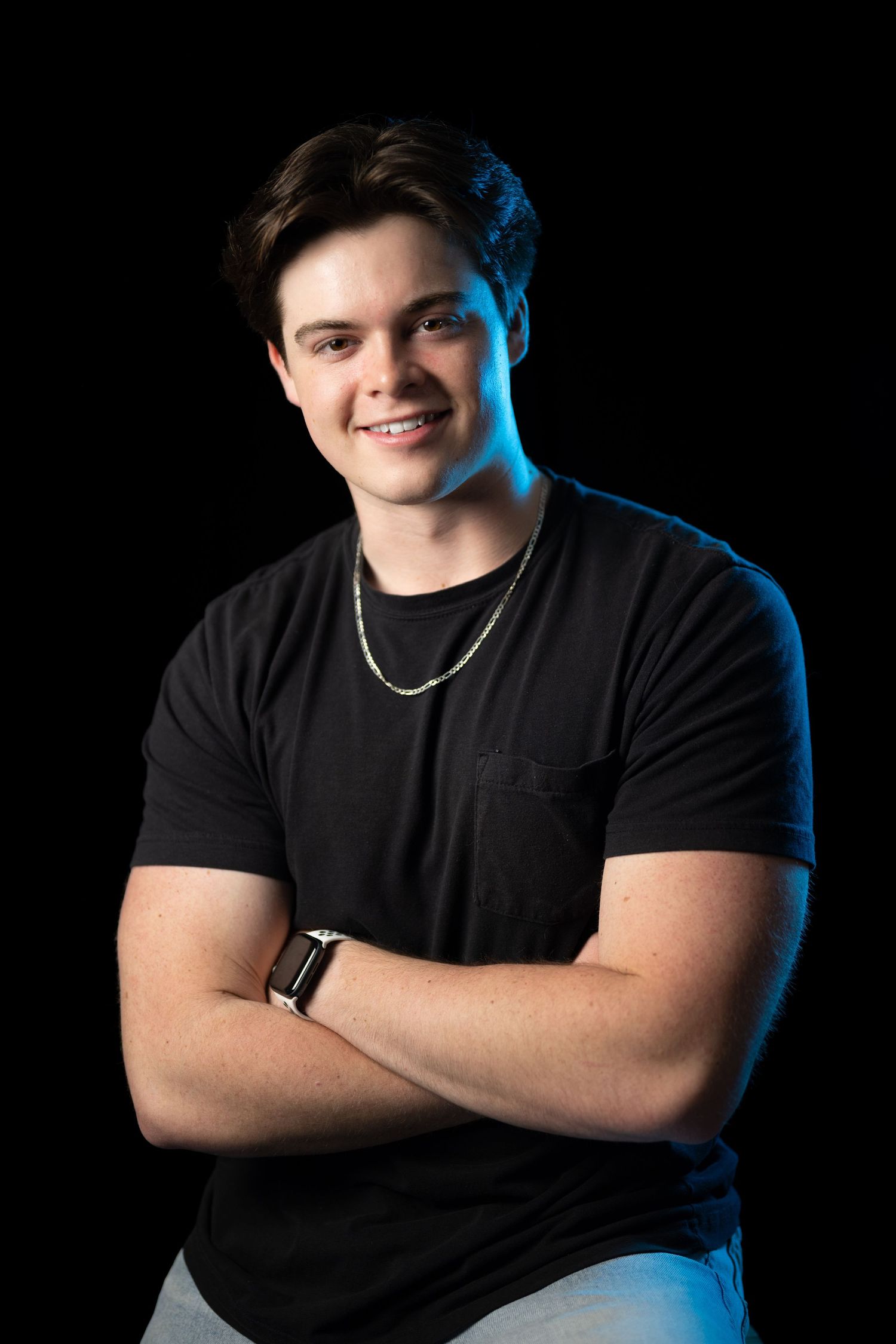 Young man in black t-shirt smiling with arms crossed against dark background with blue lighting.