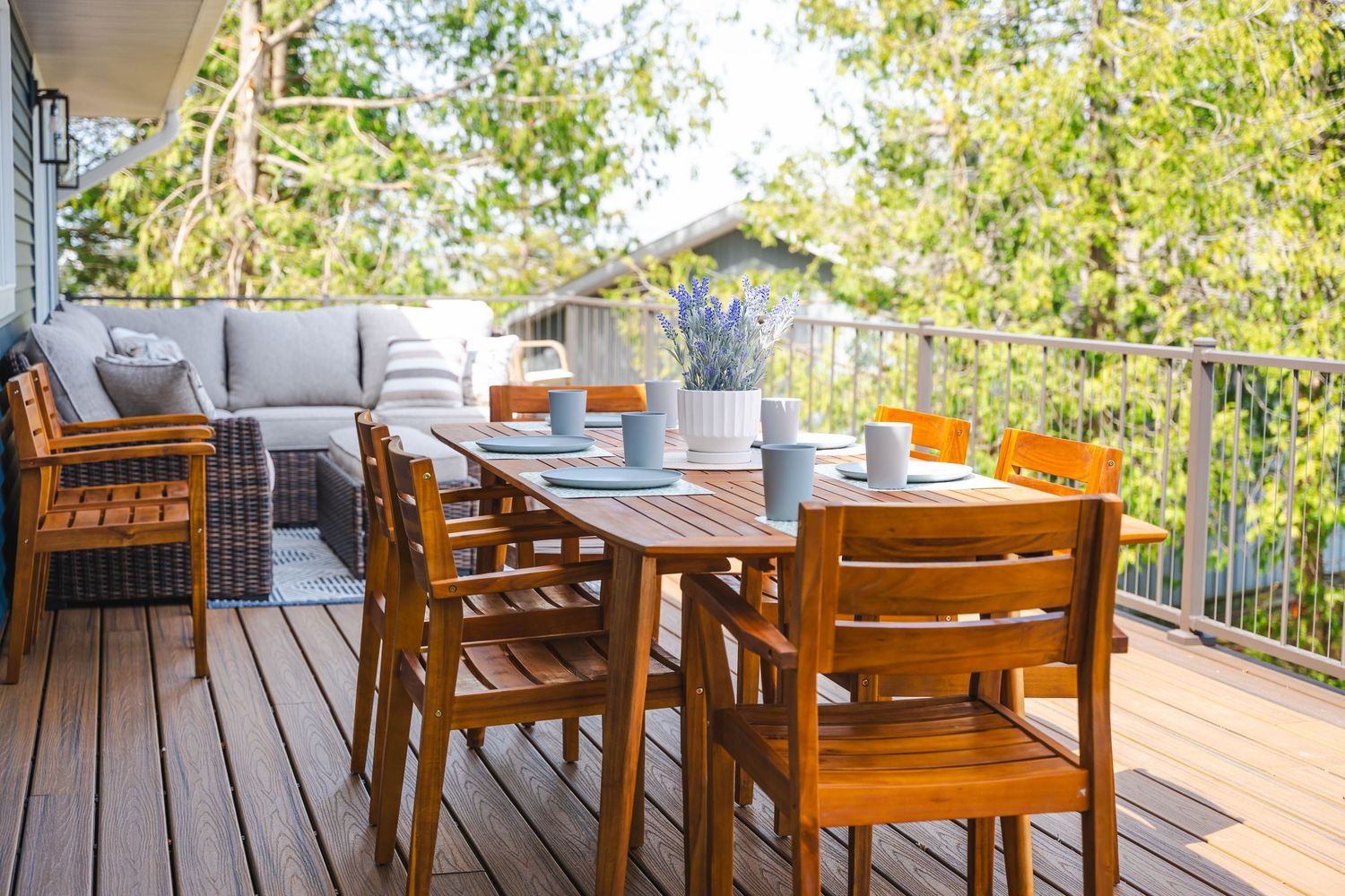 Spacious outdoor deck with wooden dining table, chairs, and sectional sofa surrounded by lush green trees.