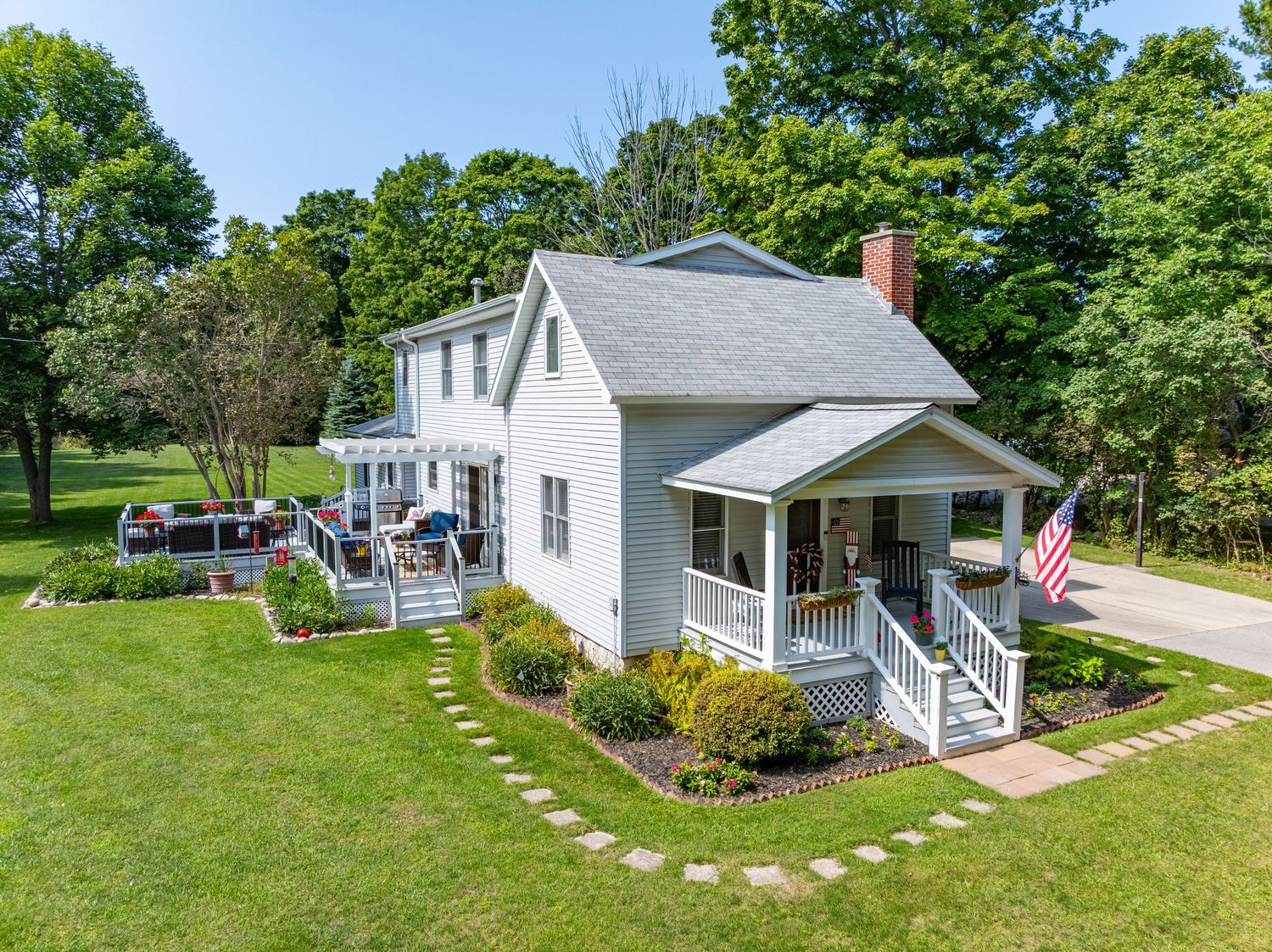 Charming white farmhouse with wraparound porch, American flag, pergola, and manicured lawn surrounded by mature trees.