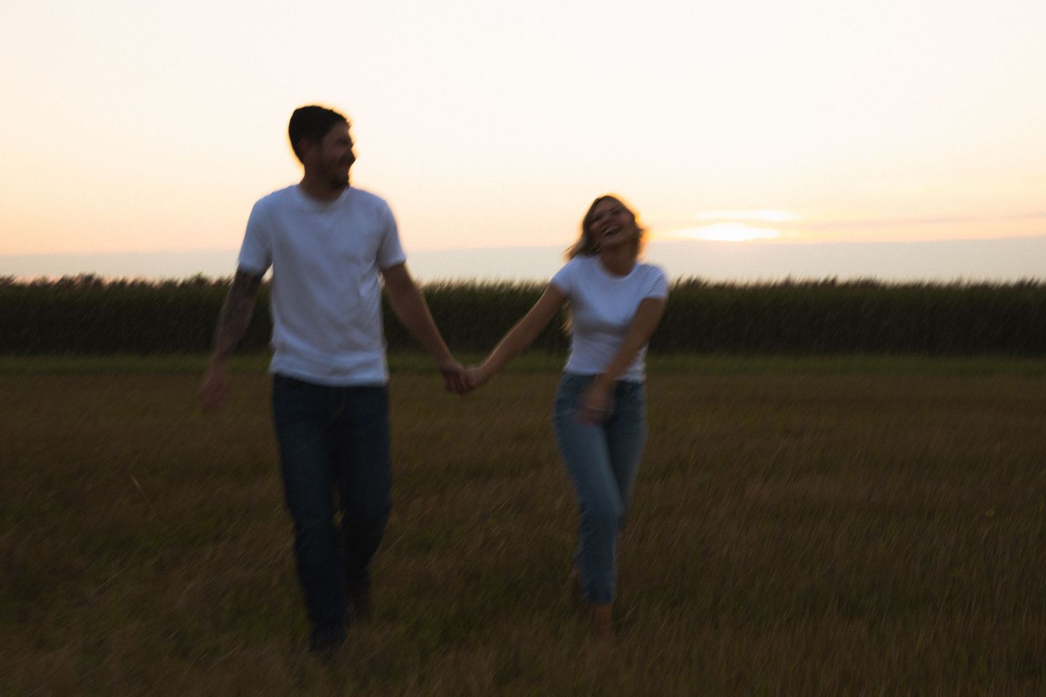 A couple in white shirts and jeans hold hands while walking through an open field at sunset.