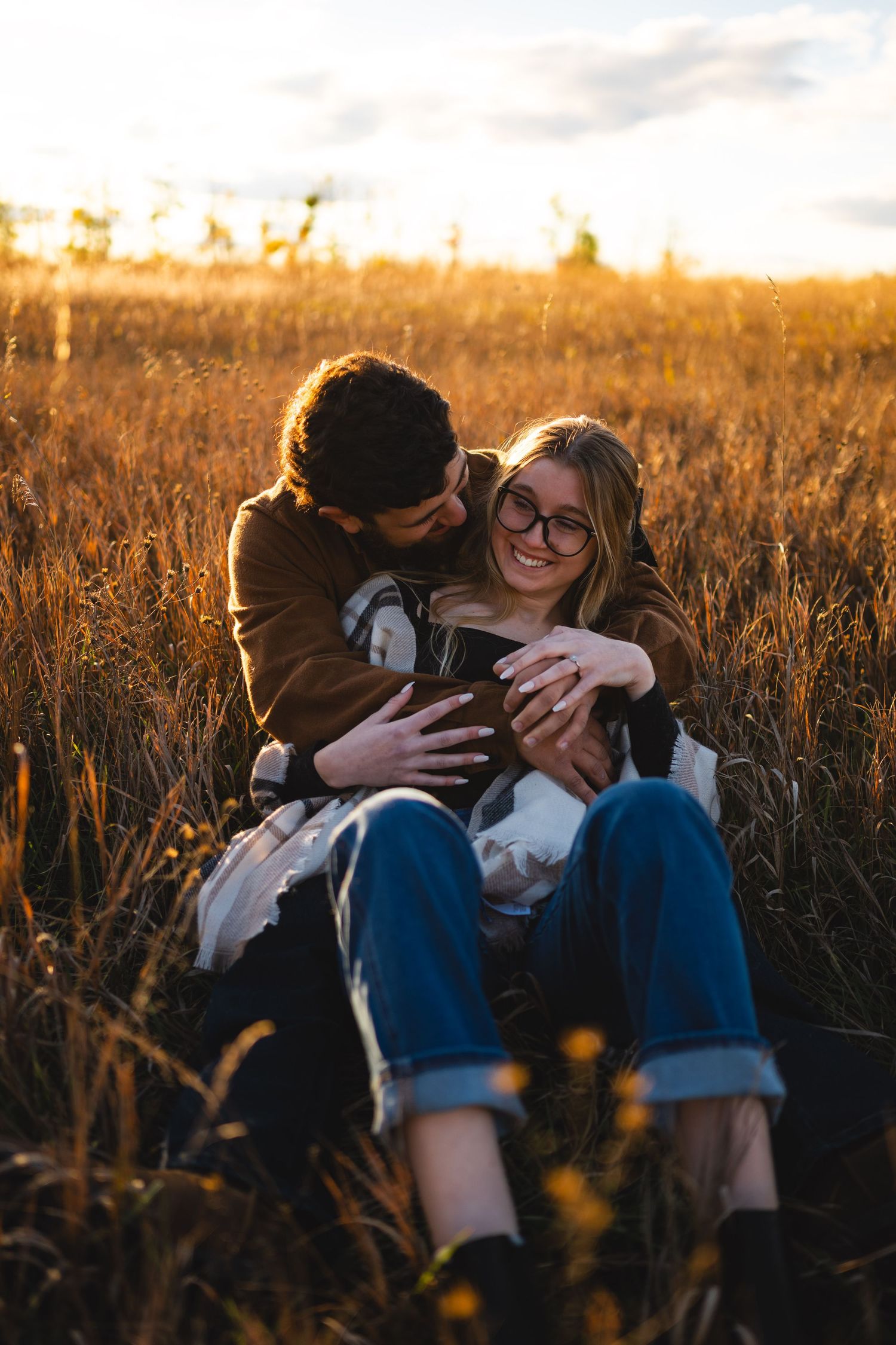 Happy couple sitting in golden autumn field at sunset, man hugging woman from behind as she laughs joyfully.