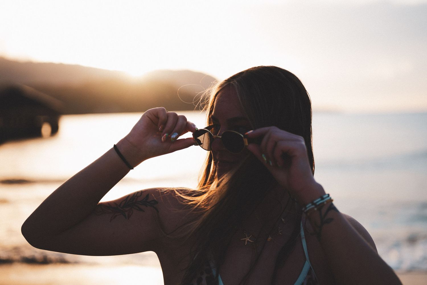 Woman adjusting sunglasses on a beach at golden sunset with tattoo visible on her arm.