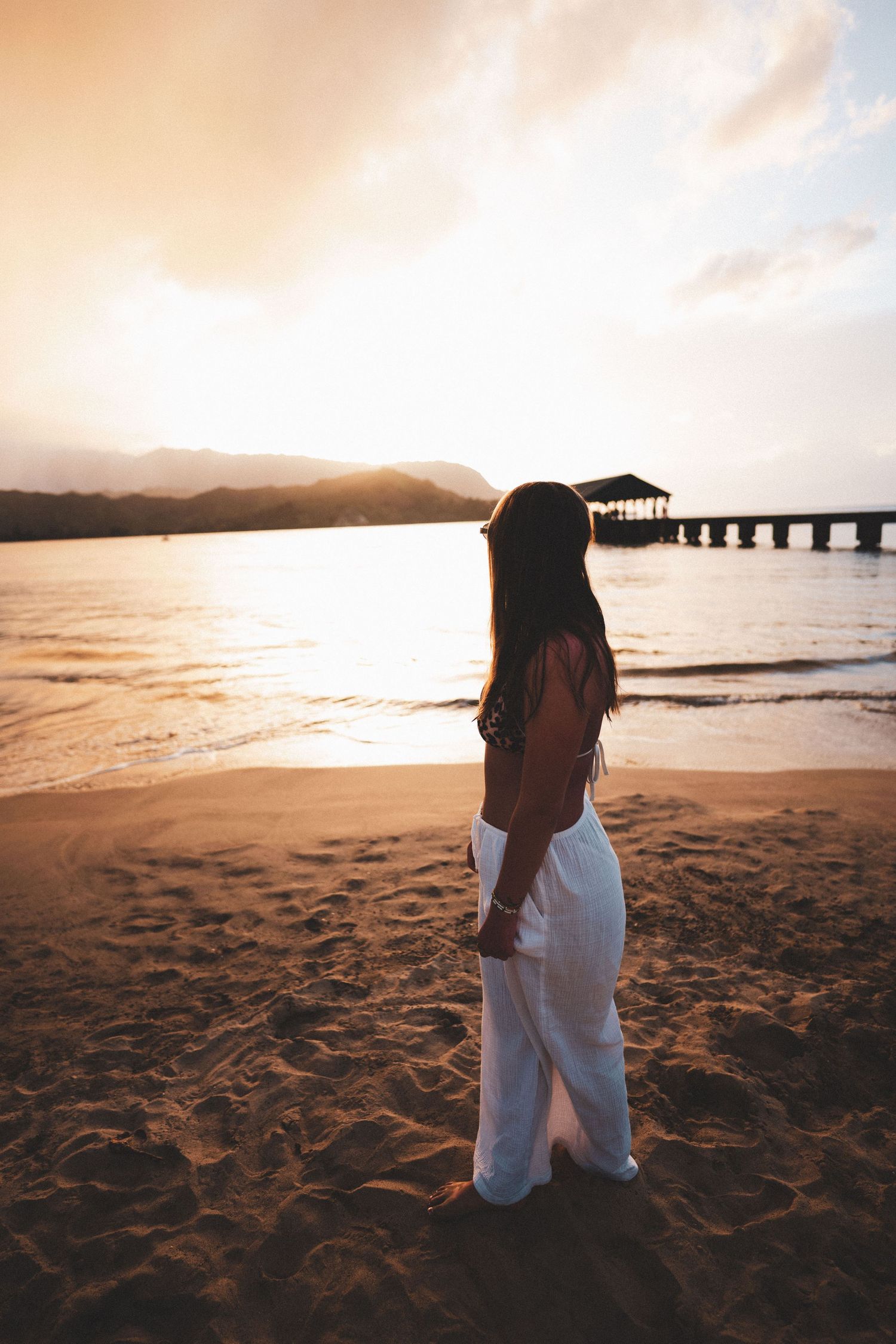 Woman in white pants standing on sandy beach at golden sunset with pier and mountains in background.