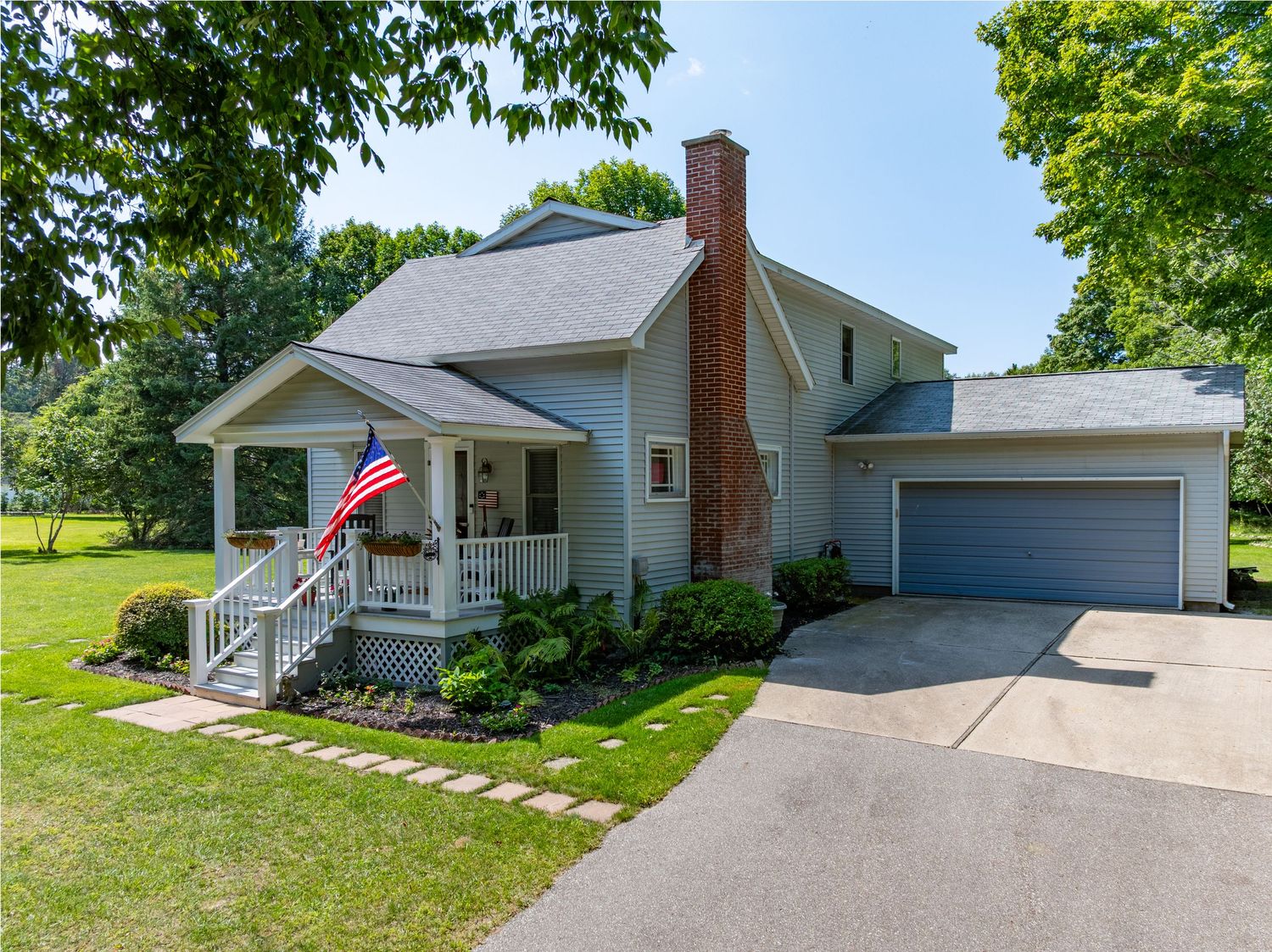 Classic American home with brick chimney, covered porch, attached garage, and American flag on a sunny summer day.