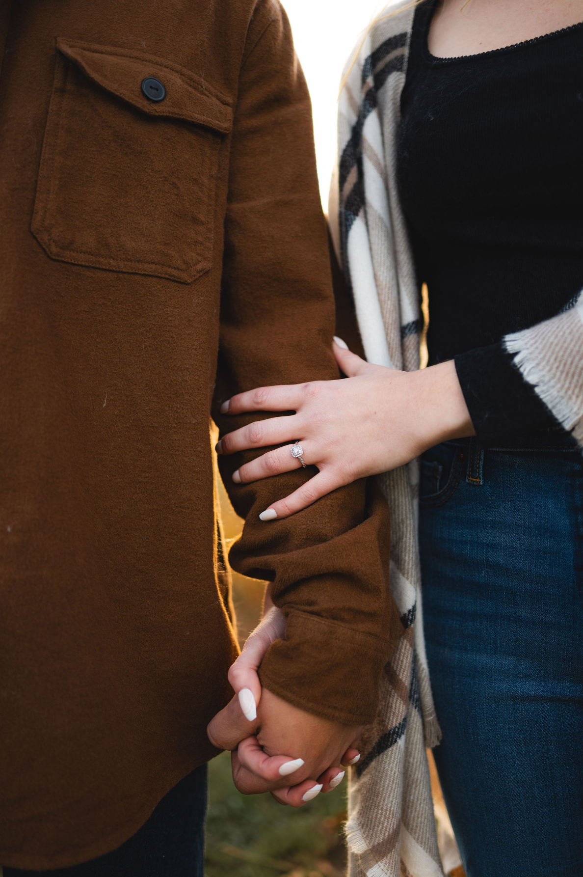 Close-up of couples hands holding, woman displaying engagement ring on mans brown jacket sleeve.