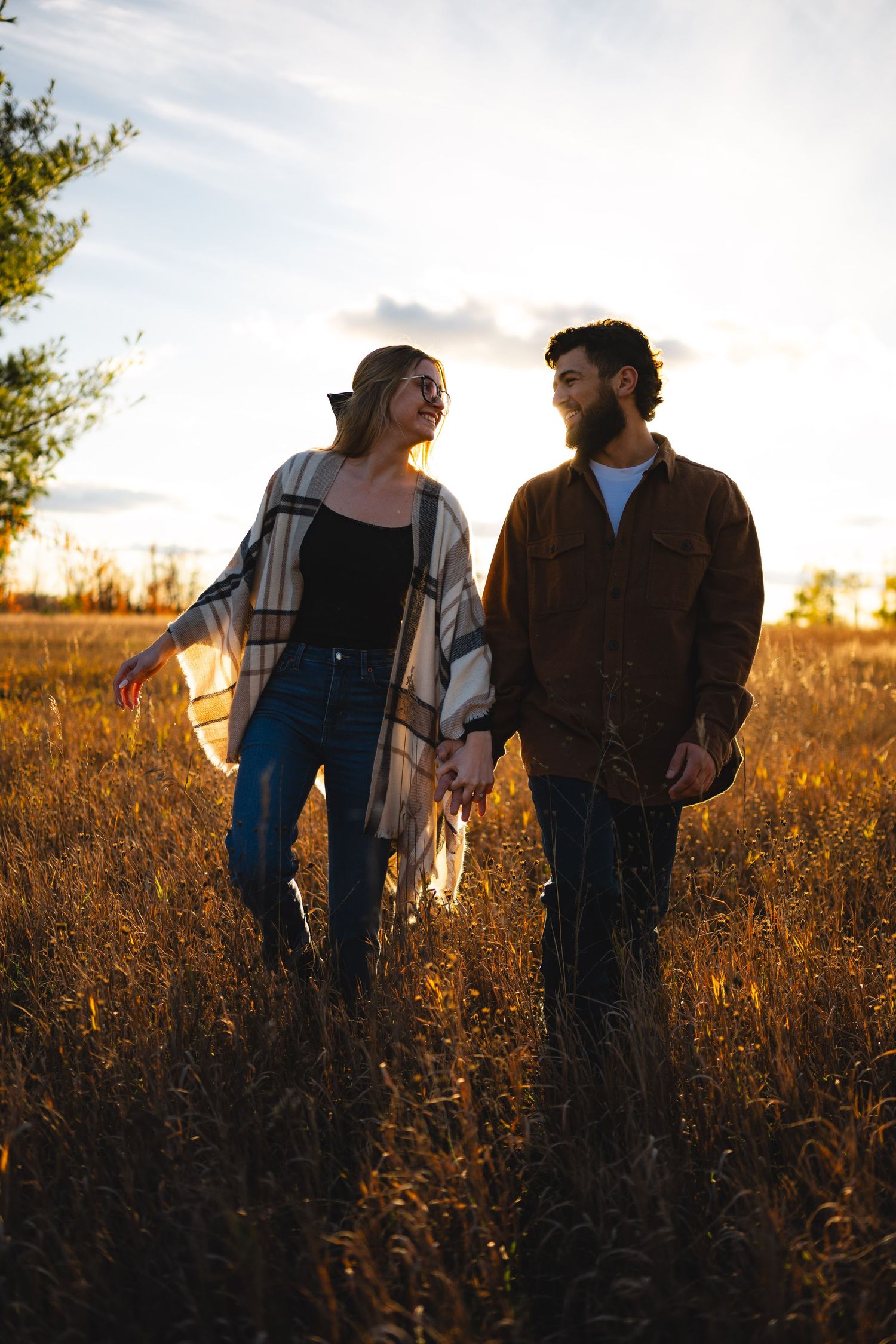 Couple holding hands walking through golden field at sunset, wearing casual autumn outfits in warm evening light.