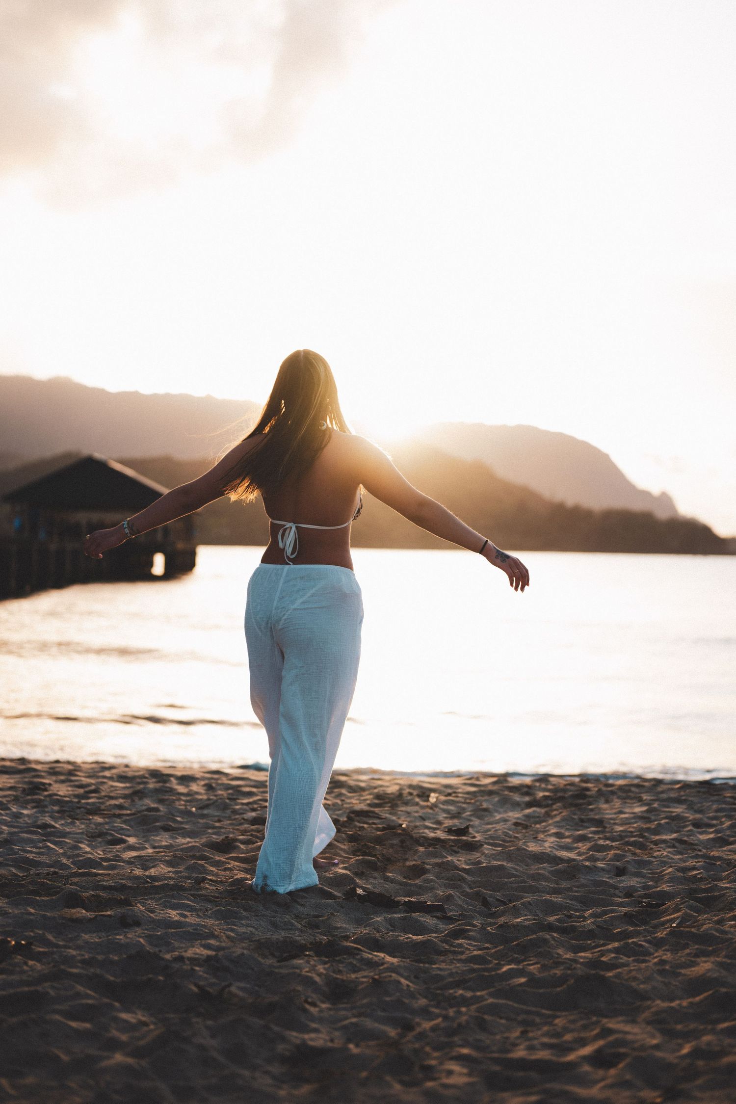 Woman in white pants and bikini top stands on sandy beach with arms outstretched at golden sunset.