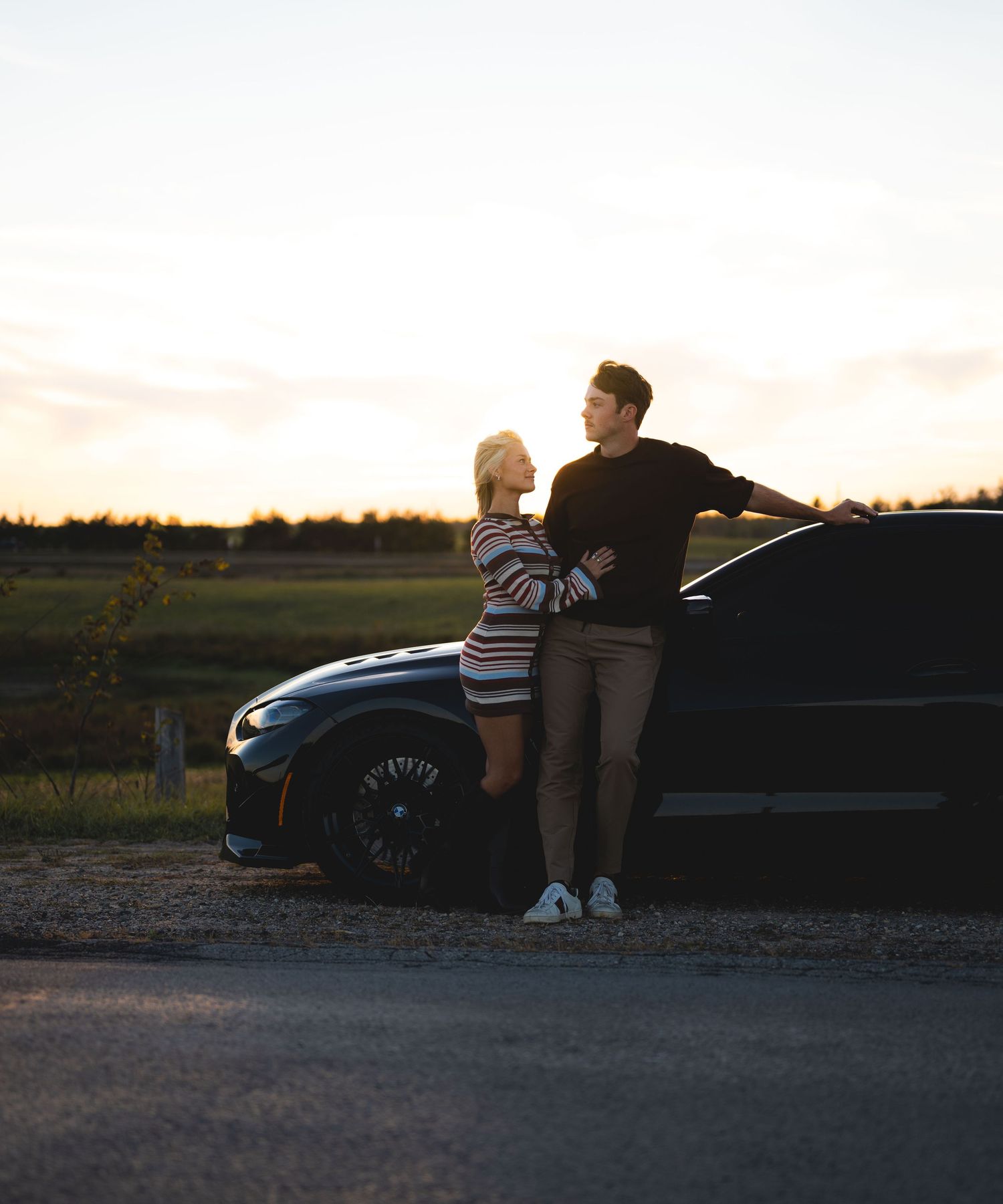 Couple leaning against a black car sharing a tender moment during a beautiful sunset.