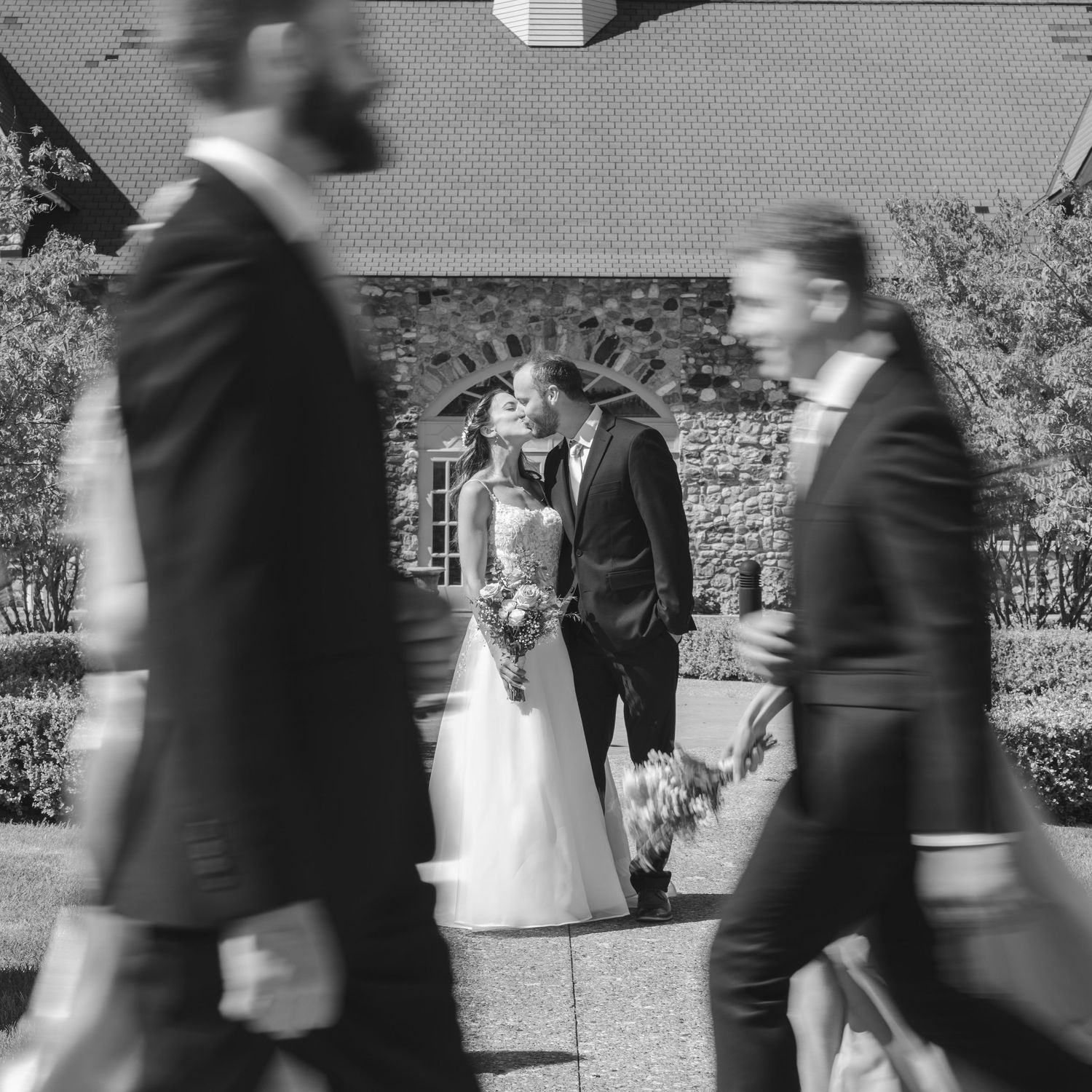 Black and white wedding photo of bride and groom kissing while guests walk past a church.