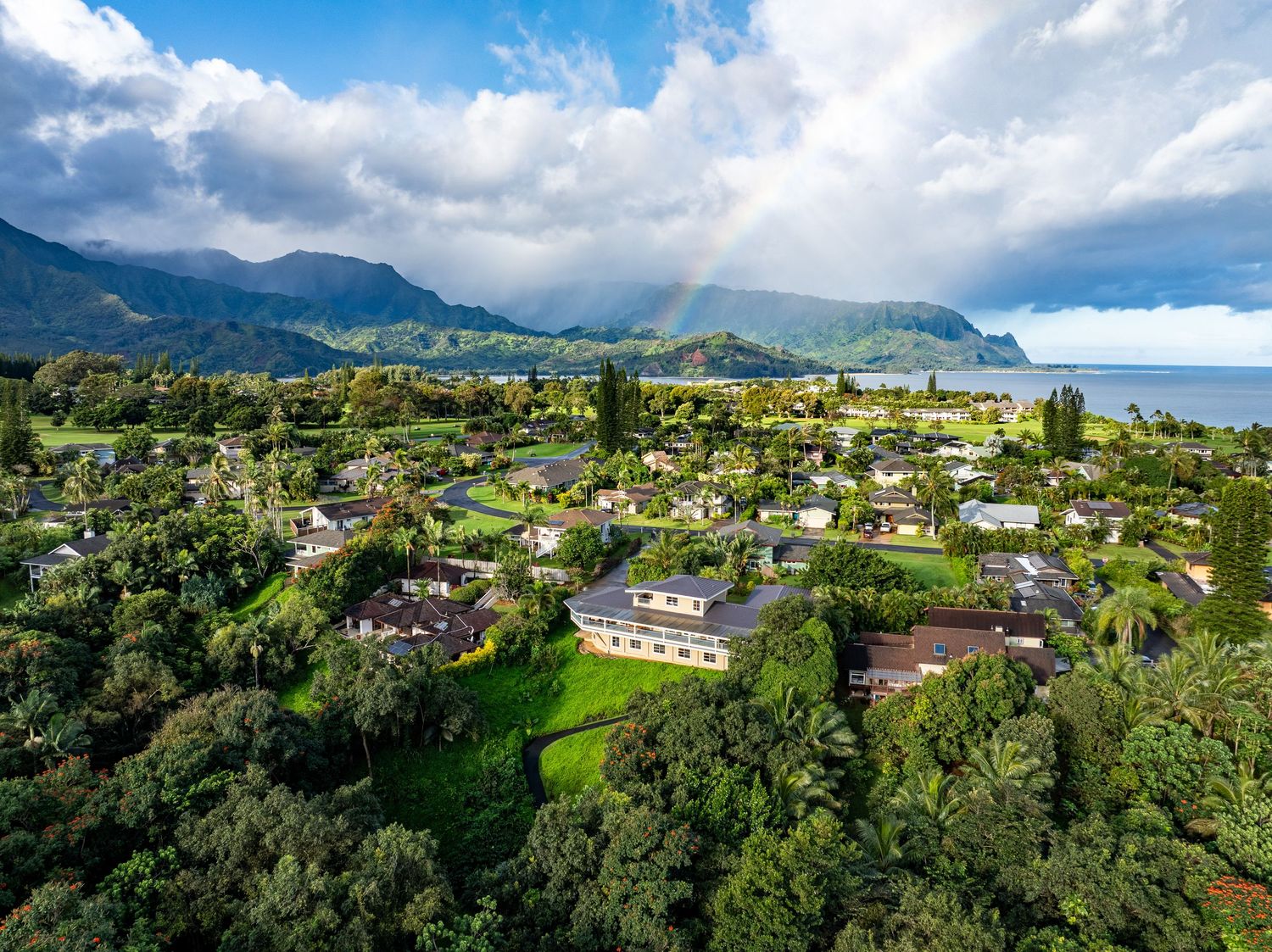 Aerial drone view of a verdant Hawaiian neighborhood with rainbow, mountain cliffs, and blue ocean in the background.