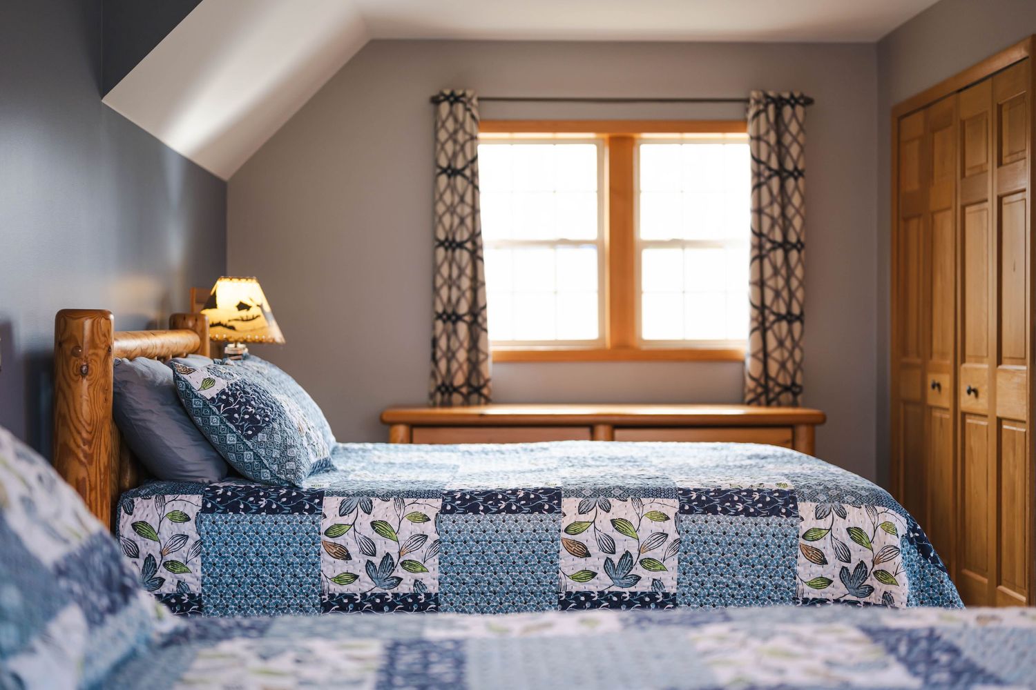 Charming attic bedroom with blue patchwork quilt, wood headboard, and dormer window.