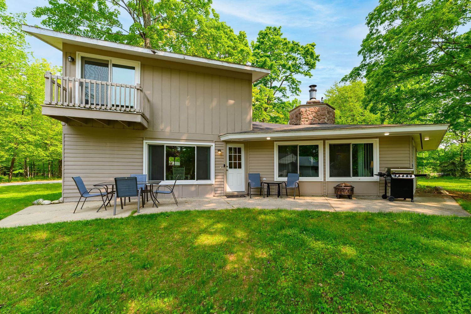 Two-story suburban home with large patio, outdoor seating, grill, and lush green lawn surrounded by mature trees.