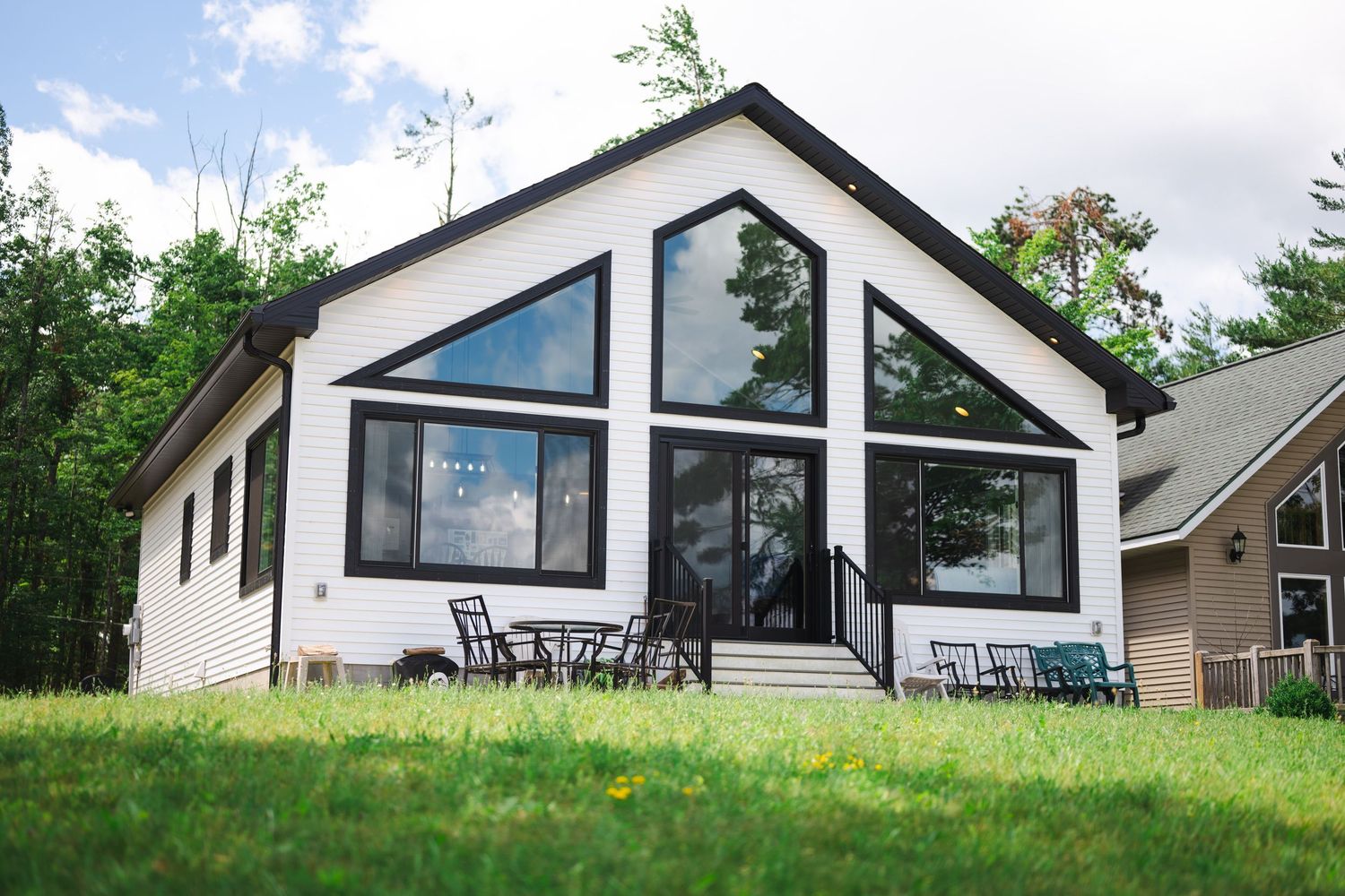 White chalet-style home with large triangular windows, outdoor seating, and green lawn on sunny day.