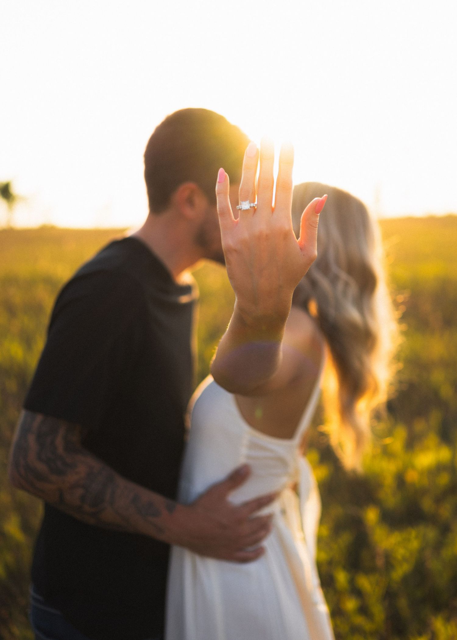 Engaged couple kissing in golden sunset field, woman displaying sparkling engagement ring toward camera proudly.