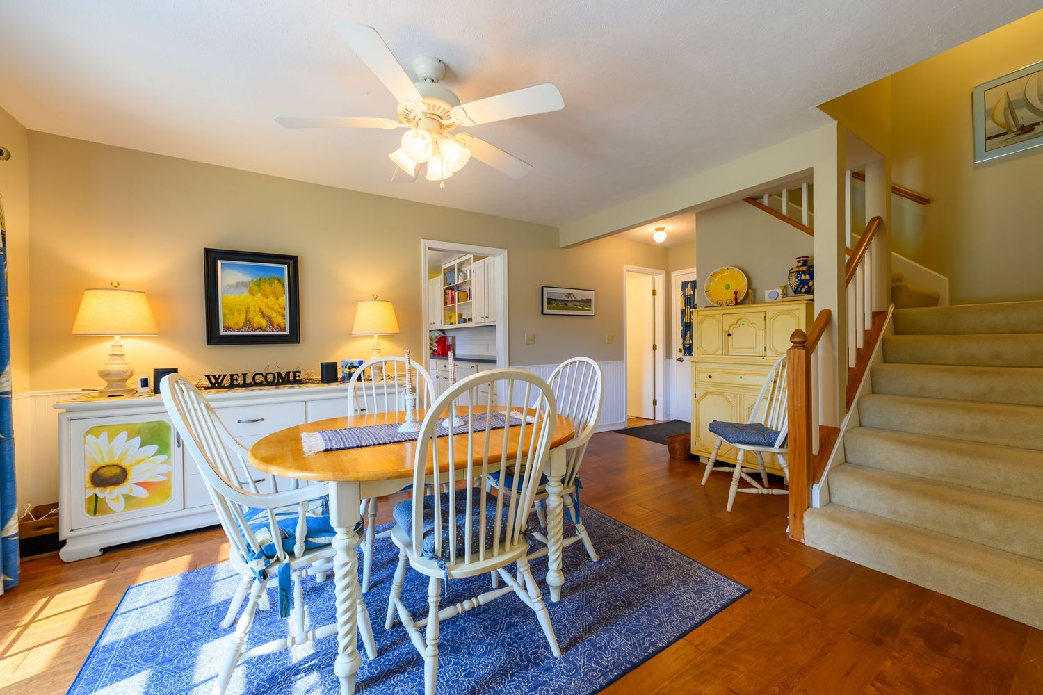 Cozy dining room with wood table, white chairs, blue rug, ceiling fan, and staircase.