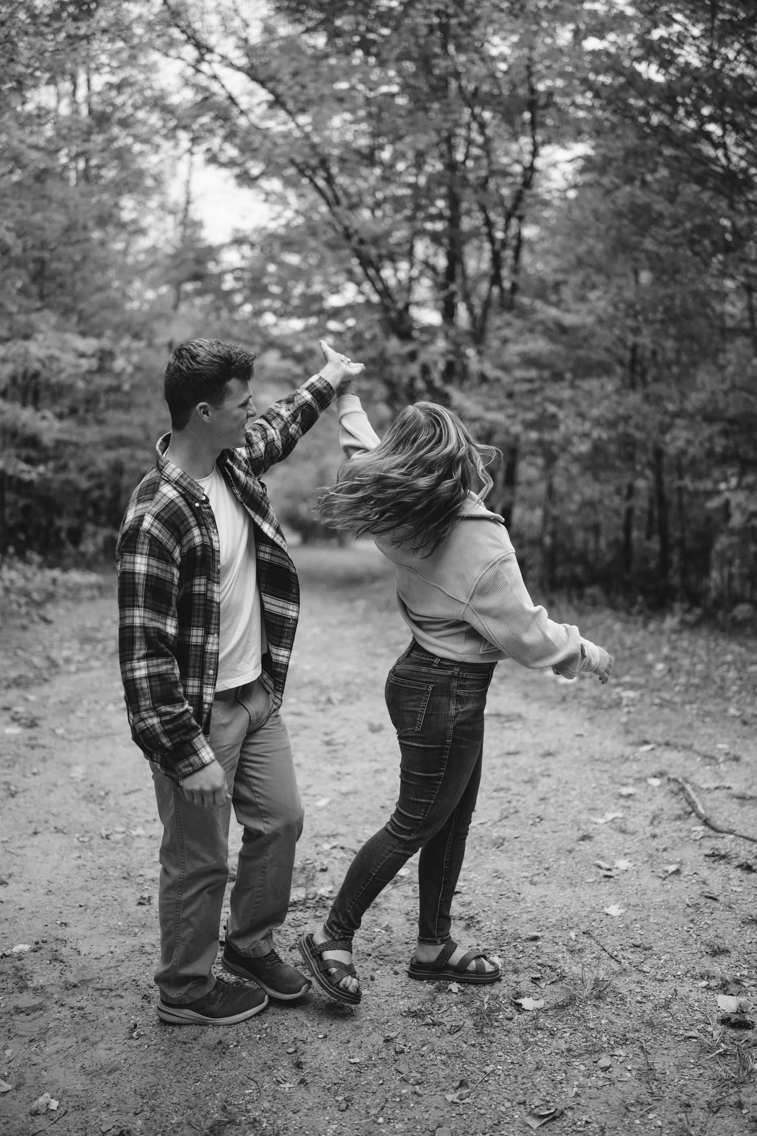 A black and white photo of a couple dancing and spinning together on a wooded forest trail in autumn.