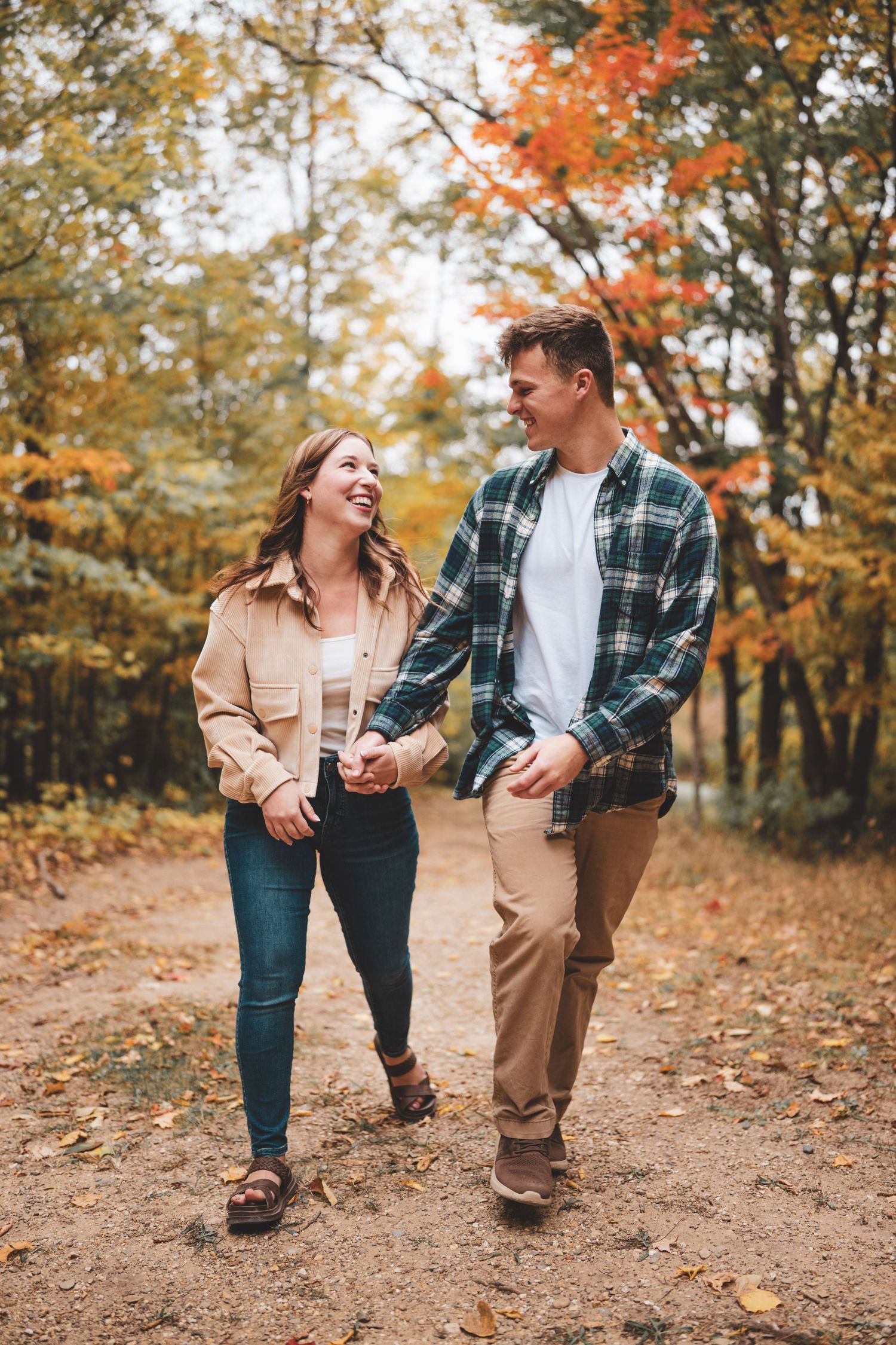 A smiling couple holds hands and walks along a leaf-covered path surrounded by vibrant autumn foliage.