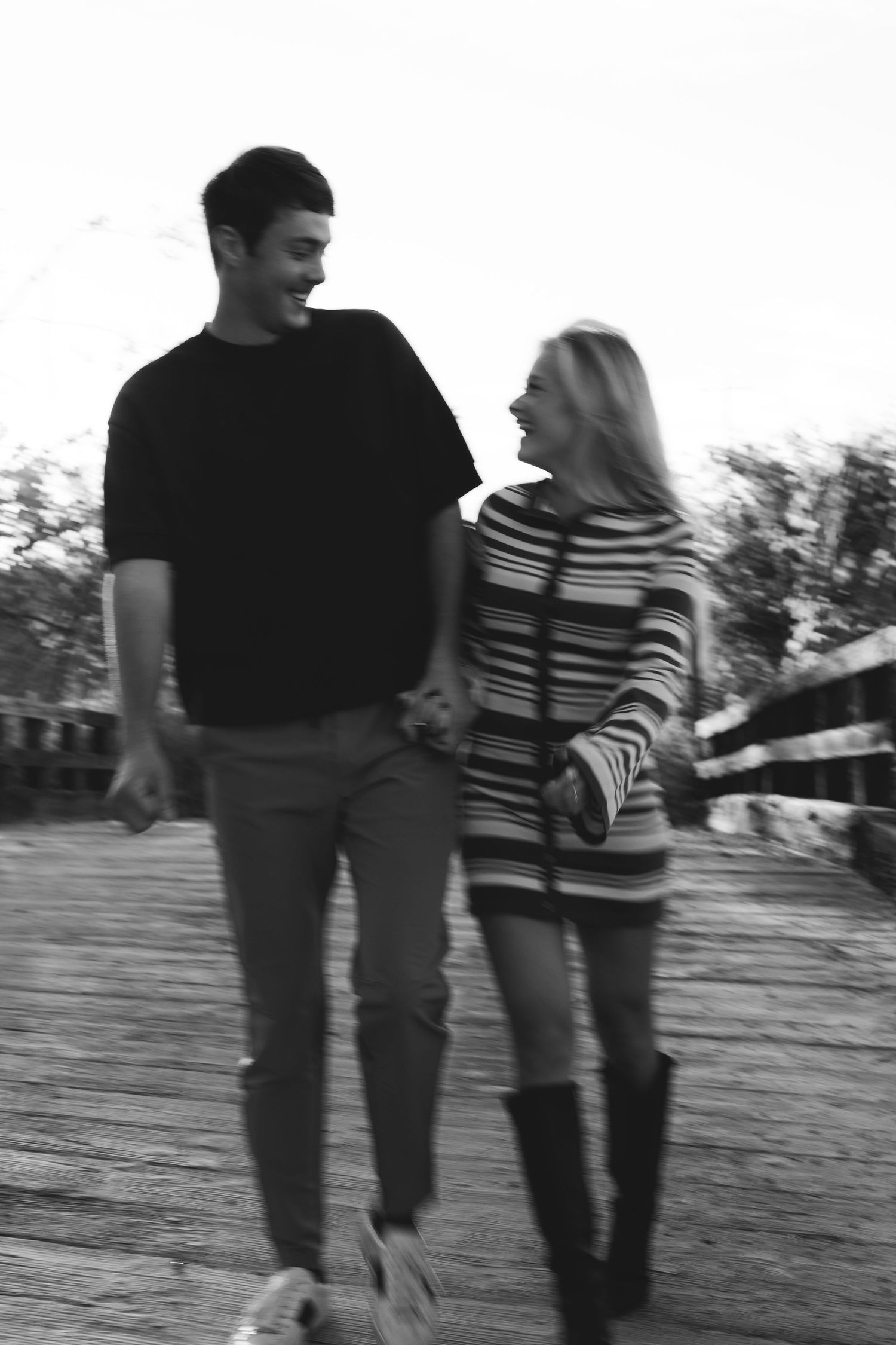 Black and white photo of a smiling couple walking hand in hand on a wooden bridge.