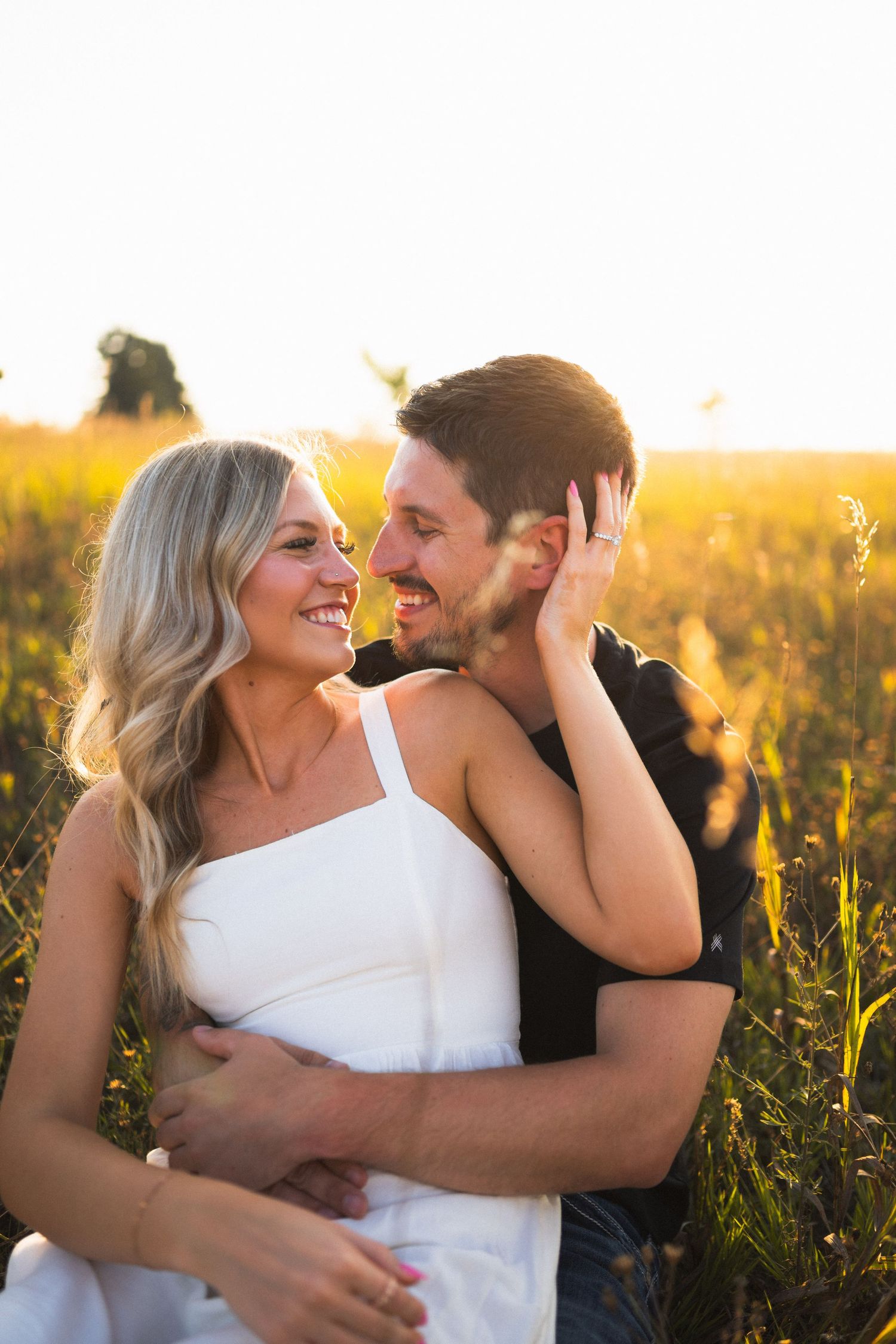 Smiling couple embracing in sunlit golden field, blonde woman in white dress laughing with man in black shirt.