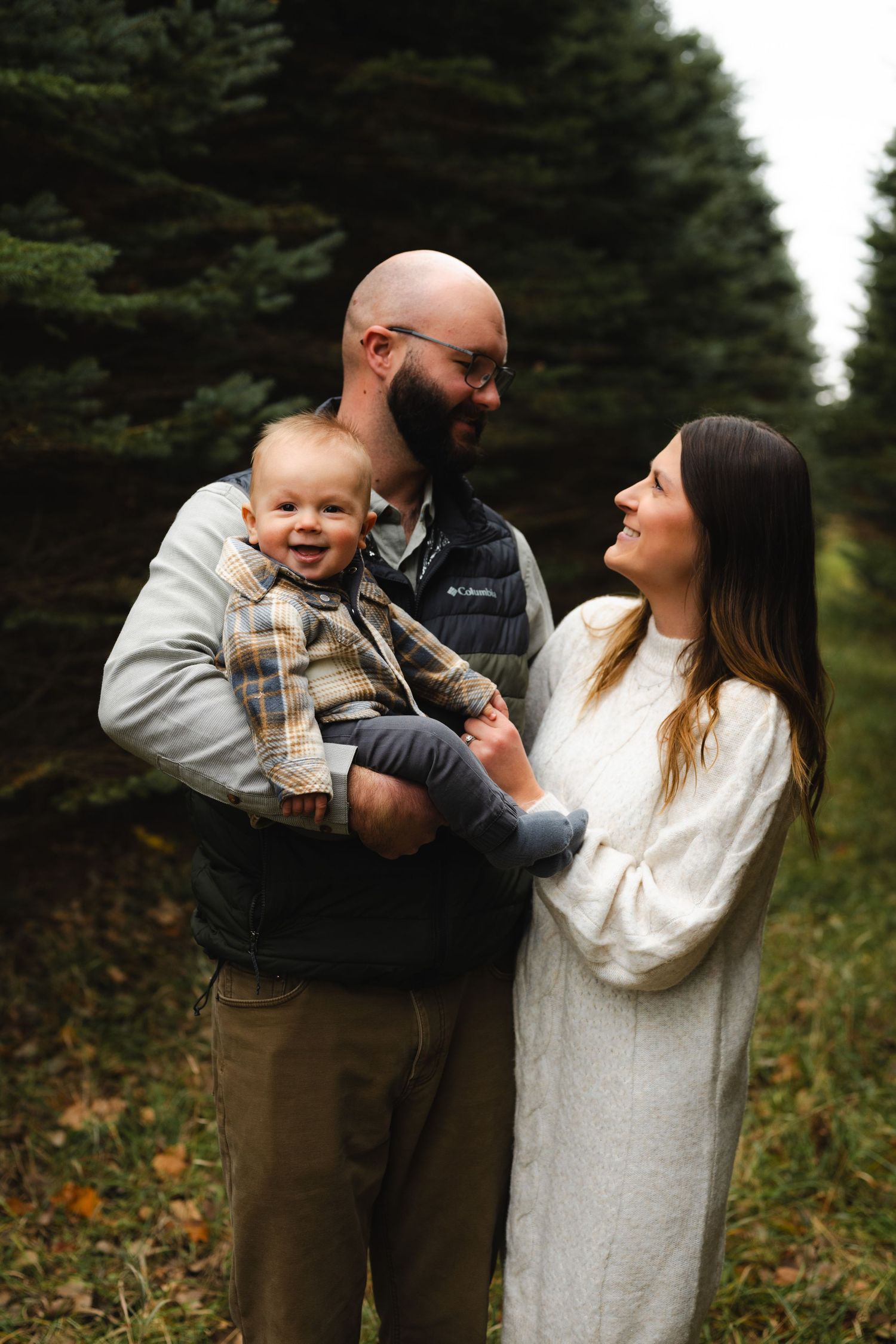 A happy family with a bald bearded dad holding a smiling baby beside a brunette mom in a white sweater among pine trees.