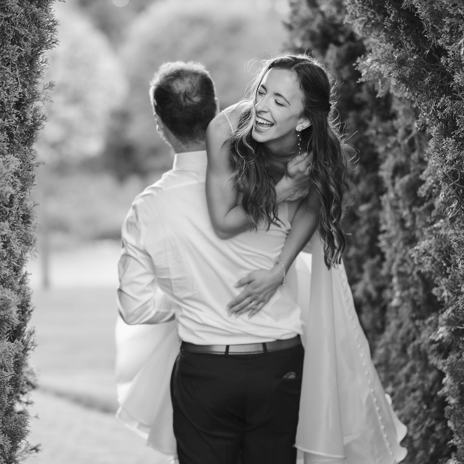 Black and white photo of groom lifting laughing bride between tall hedges in a garden.