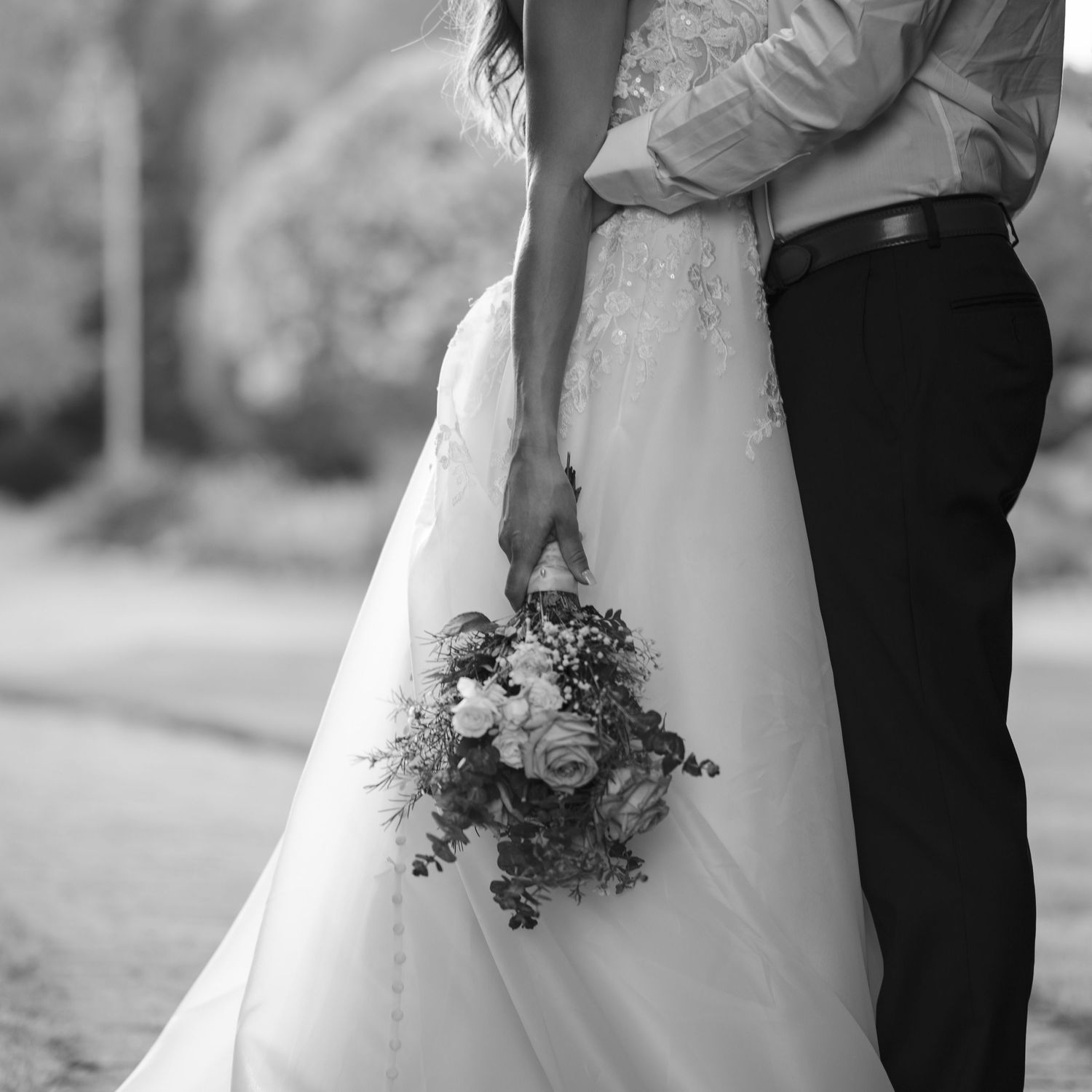 Black and white wedding photo of couple embracing, bride holding a bouquet of roses in flowing gown.