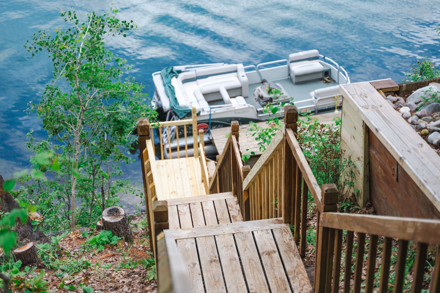 Wooden staircase leading down to a lakeside dock with a pontoon boat on calm blue water.