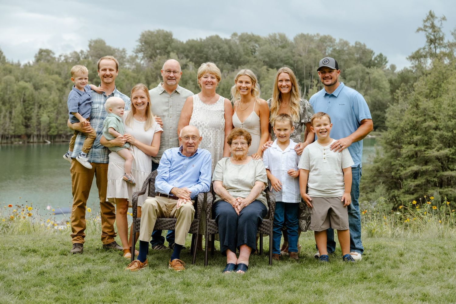 Multi-generational family portrait of 11 people smiling outdoors near a lake surrounded by lush green trees.