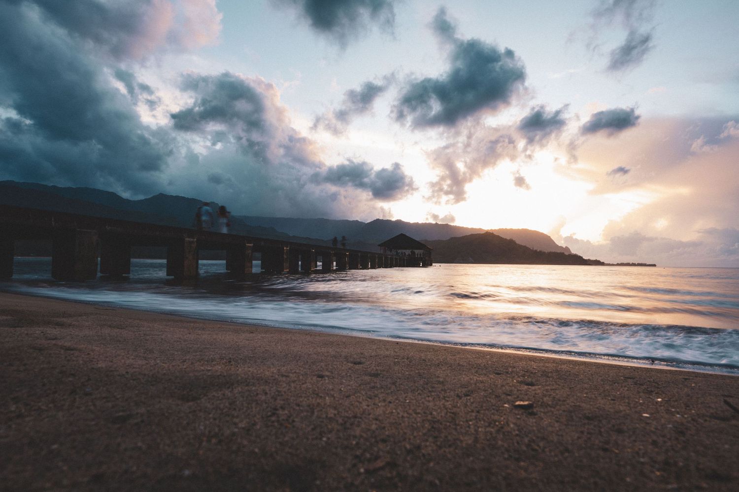 Dramatic sunset over a sandy beach with a historic pier building and mountain silhouette under cloudy skies.