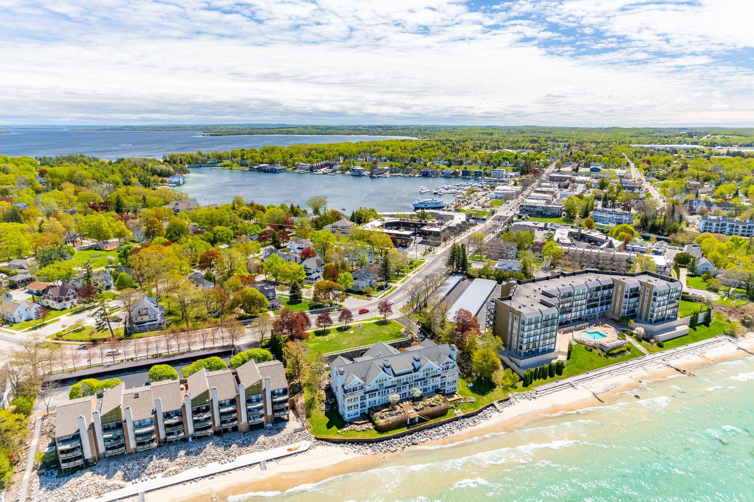 Aerial view of a lakeside town with condos, marina, and sandy beach along bright blue waters on a sunny day.