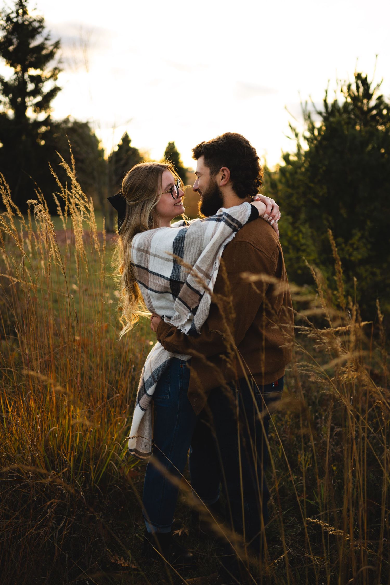 Romantic couple nearly kissing in tall grass field at sunset, woman in plaid wrap and jeans embracing bearded man.