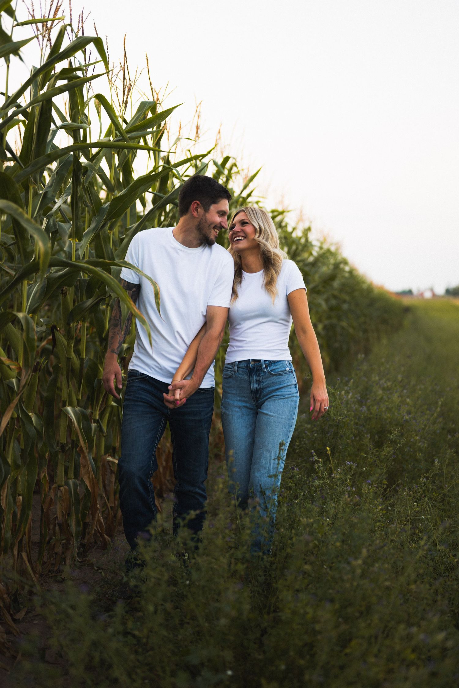 A couple in matching white shirts and jeans hold hands and smile at each other while walking beside a lush cornfield.