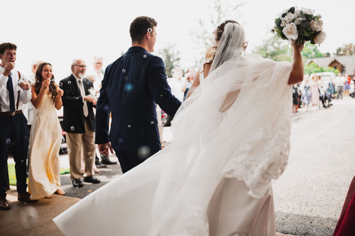 Bride and groom walking away after wedding ceremony as guests celebrate with bubbles outdoors.